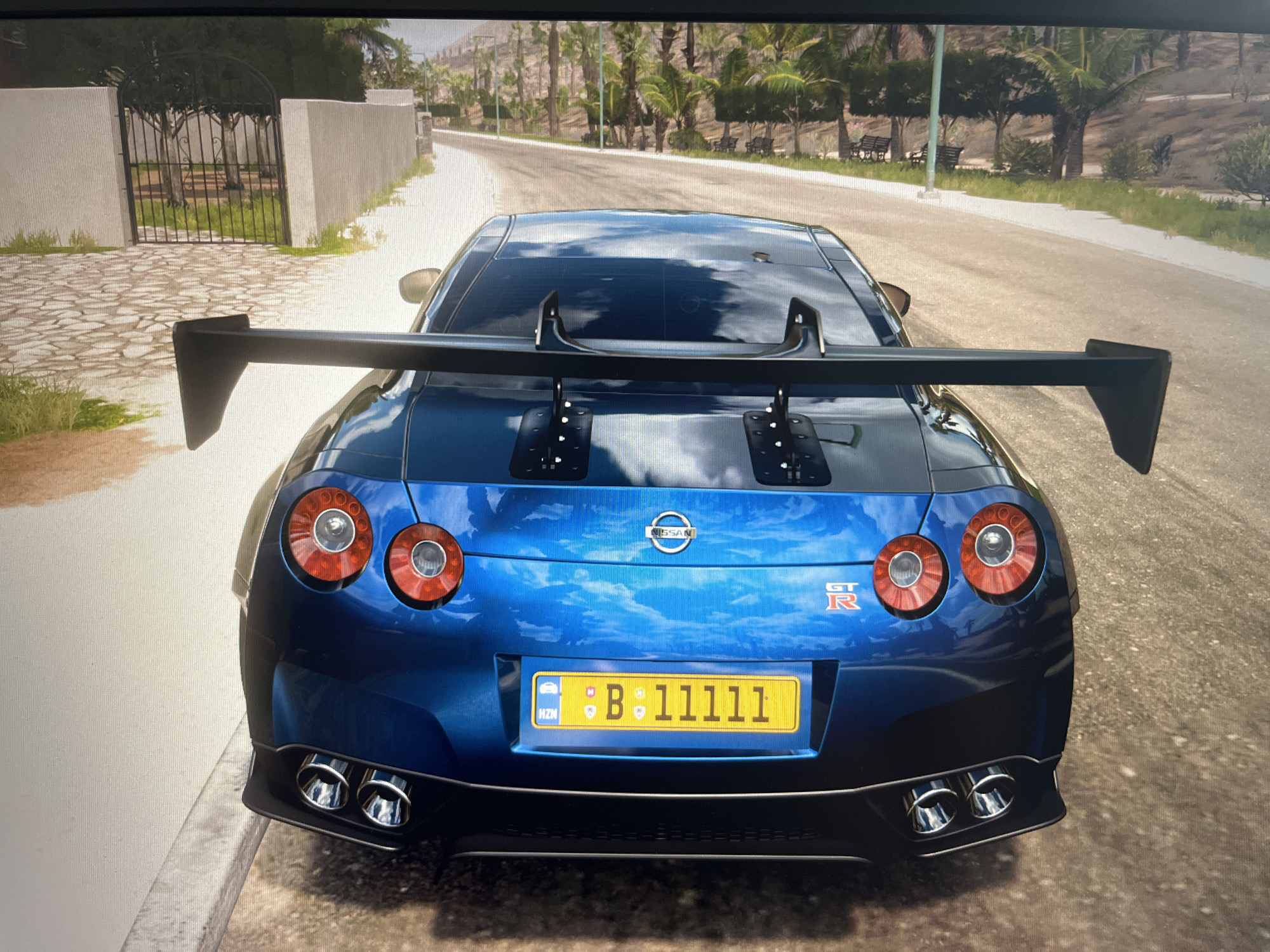 The image shows the rear view of a blue Nissan GT-R with a large spoiler, parked on a road in a sunny, tropical-like environment. The car has a yellow license plate with the characters "B: 11111" and what appears to be a Romanian or Bulgarian designation "HZM" on the left. The background features palm trees, a paved road curving to the left, and a white wall with a black metal gate on the left side of the image. There are no people visible in the image. The scene depicts a car in a video game, likely Forza Horizon, given the graphical style and user interface elements like the license plate.