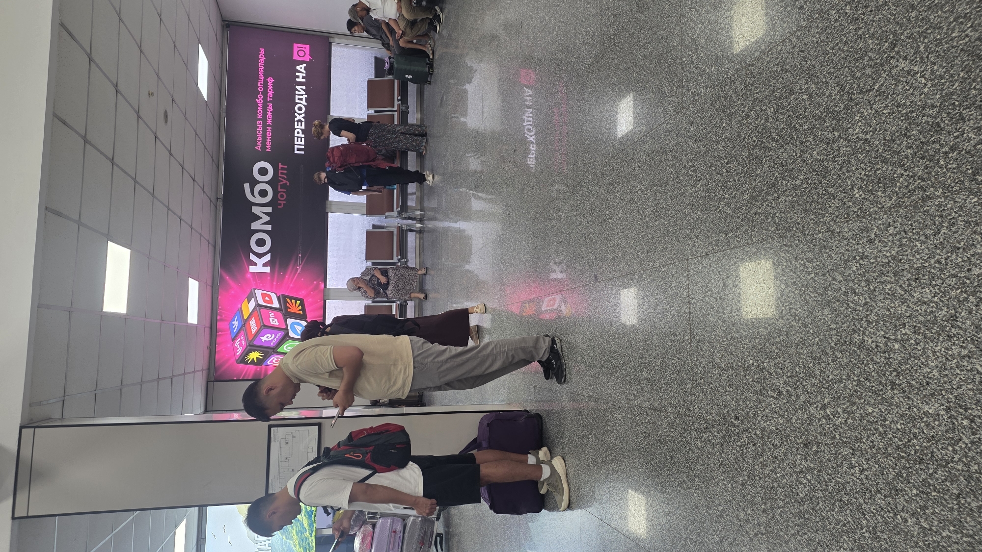 The image depicts a scene inside an airport terminal. In the foreground, two men are standing, looking at their phones. The man on the left wears a white t-shirt and black shorts, with a red backpack on his back. The man on the right wears a beige t-shirt and gray pants. A purple duffel bag is on the floor next to the man on the left. Behind them, a large advertisement is displayed on the wall, featuring colorful icons and the text "КОМБО" in large letters. Several other people are visible in the background, seated on benches or standing. The floor is tiled and reflective. The environment suggests a public waiting area, likely at an airport, due to the presence of seating, an advertisement for a service, and people with luggage. The location is identified as Nariman, Kyrgyzstan.