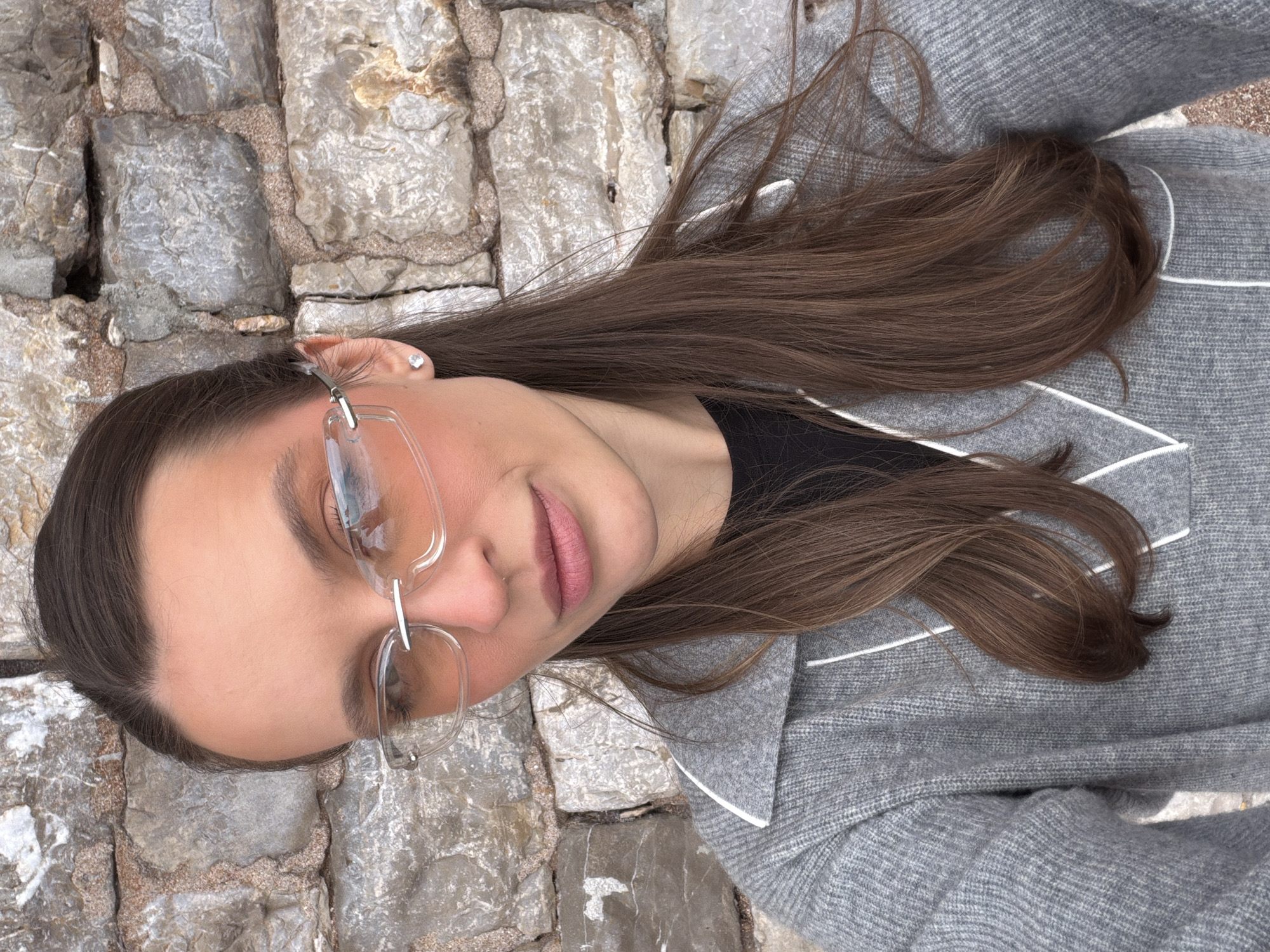 A young woman lies on a stone surface in Petrovac centar, Montenegro. She wears glasses with clear frames and a gray sweater with white piping. Her long, brown hair is partially spread out on the stones. She has light skin, pink lips, and a silver heart-shaped earring in her left ear. The stone surface appears to be a path or ground made of irregularly shaped, gray stones.