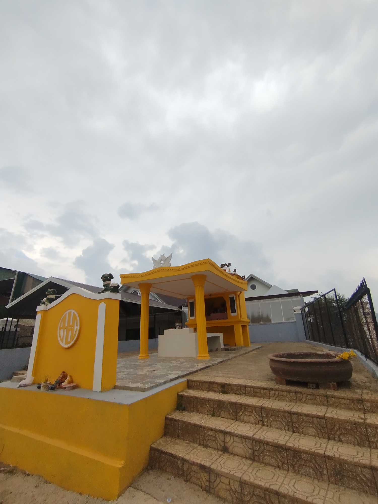 A yellow structure with a peaked roof and white trim is at the center of the image. The structure is surrounded by a concrete platform and stairs. The sky is cloudy and gray. The location is in the city of Lâm Viên - Đà Lạt, Vietnam.