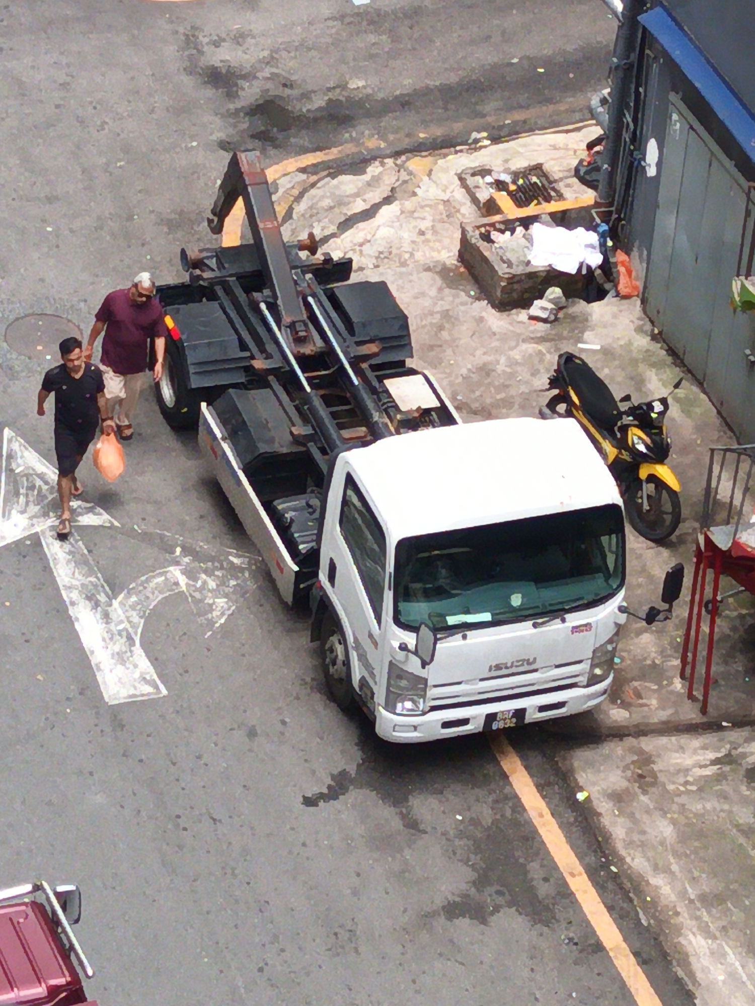 An overhead view captures a street scene in Kuala Lumpur, Malaysia, where a white Isuzu flatbed truck is parked. The truck's rear mechanism is raised, with its hydraulic arms extended. A yellow and black motorcycle is parked to the right of the truck. Two men are walking away from the truck on the left side of the frame. The man in the foreground wears a black t-shirt and shorts, carrying an orange bag. The man behind him is wearing a maroon t-shirt and light-colored shorts. The ground is asphalt, with white directional arrows painted on it. In the background, a building with a gray facade and blue trim is visible, along with some stacked crates and debris.