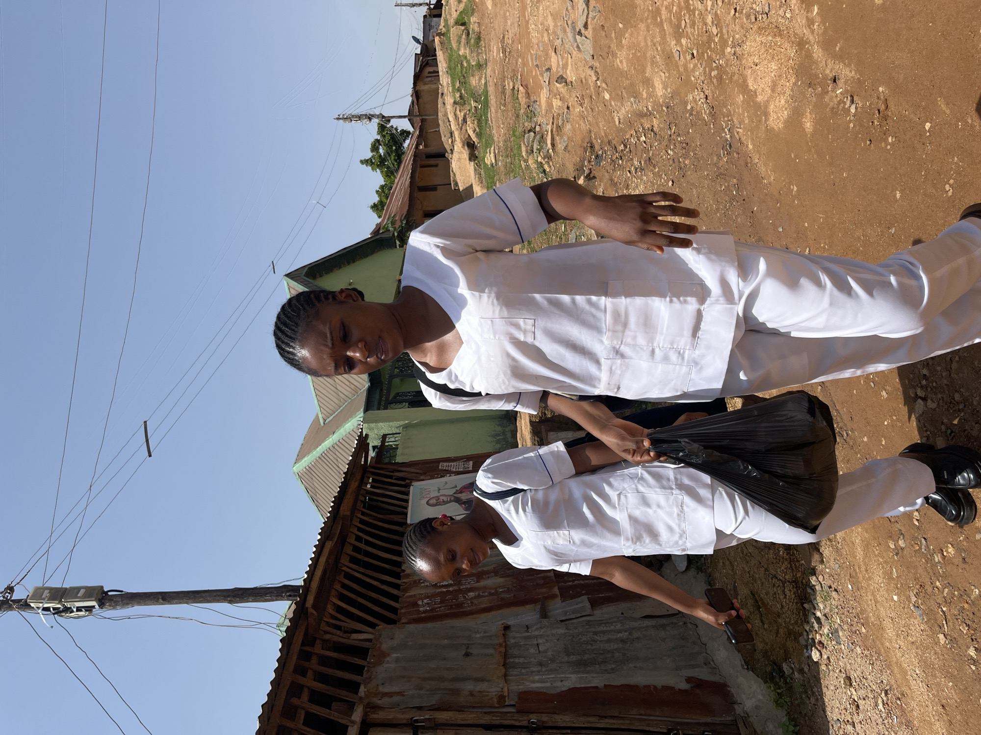Two young women in white nurse uniforms walk down a dirt road in a residential area of Jos, Nigeria. The woman on the right is closer to the camera and is holding a black plastic bag in her right hand. The woman on the left is slightly behind and to the left of the first woman, and she is holding a smartphone in her left hand. They are both looking forward and appear to be walking together. The road is unpaved and dusty, with scattered rocks. In the background, there are several buildings with corrugated metal roofs, some of which appear to be made of mud or concrete. A wooden utility pole with wires runs along the left side of the road. The sky is clear and blue. The overall impression is of a simple, working-class neighborhood in a warm climate.