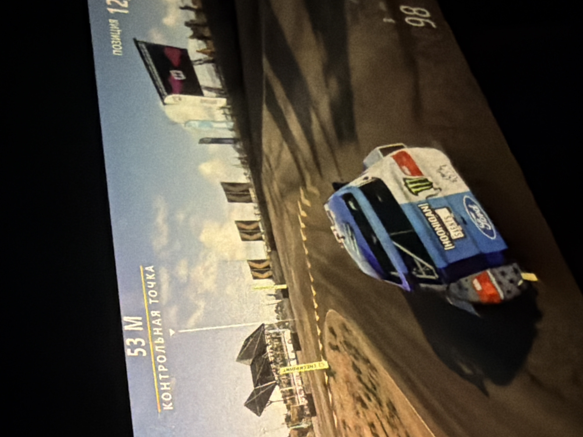 The image depicts a racing video game, likely played on a screen or monitor. The game features a blue and white rally car with "HOONIGAN!" branding and a Monster Energy logo. The car is actively driving on a dirt road circuit with visible trackside banners. The game's interface displays in-game data such as "53 M" and the word "CHECKPOINT." Also shows the text "ПОЗИЦИЯ 12", meaning "Position 12." There is an unreadable number "86" next to the car.
The location in the city of Khimki, Russia, is unknown.