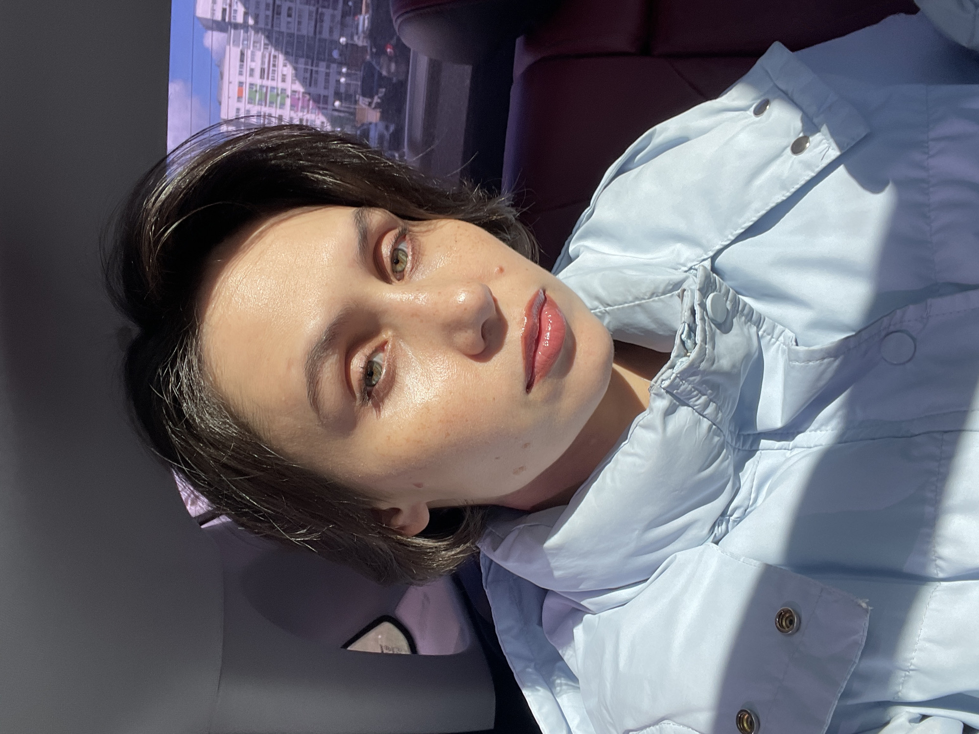 A fair-skinned woman with short, dark hair and hazel eyes poses indoors. She is wearing a light blue puffer jacket. The background shows glimpses of a city scene with buildings. The location is suggested to be Izhevsk, Russia.