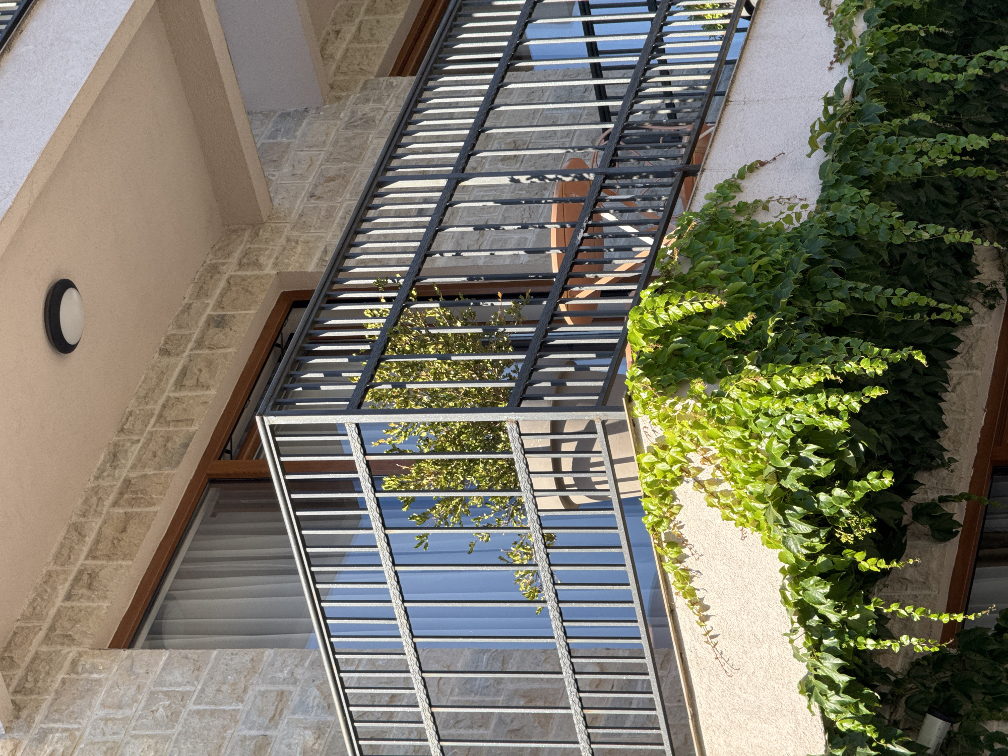 This is a close-up, eye-level shot of a balcony on a multi-story building in Budva, Montenegro. The balcony has a dark metal railing with vertical bars and a decorative wavy pattern on the top rail. A large planter containing a green leafy plant is visible behind the railing, and a small round table with chairs is also present on the balcony. The building's exterior is clad in light-colored stone blocks. Lush green ivy is growing and trailing down the side of the building beneath the balcony. A circular light fixture is mounted on the underside of the balcony above. The image is taken during daylight with clear sunlight casting shadows.