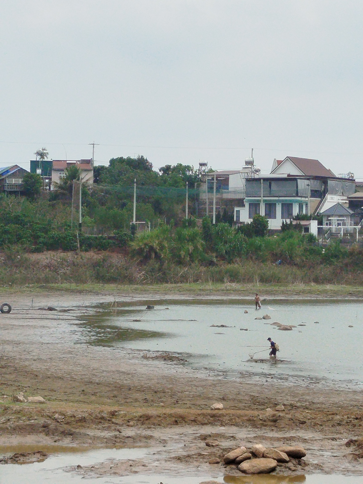 The image depicts a muddy, partially submerged landscape in Tân Đà, Vietnam, with a background of houses and greenery on a slight rise. Two people are wading in the shallow water, one in the distance and one closer to the foreground, both appearing to be fishing with nets. A discarded tire is partially submerged near the left edge of the frame. The sky is overcast. The foreground is characterized by muddy terrain and scattered rocks.