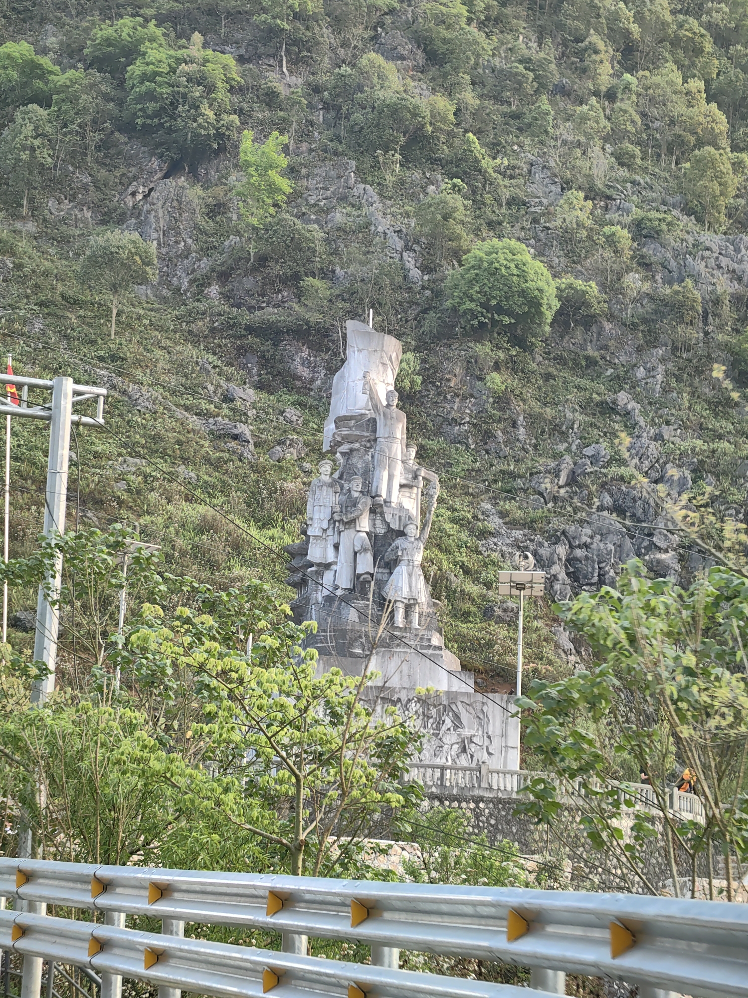 The image depicts a large, monumental statue in Đồng Văn Commune, Vietnam, situated against a backdrop of a rocky, tree-covered hillside. The statue is composed of several figures and elements stacked vertically, appearing to be constructed from a light-colored stone. Below the figures, a sculpted frieze or relief panel appears along a wall. The scene is partially obscured by foliage in the foreground, with a metal guardrail visible at the very bottom of the image. Utility poles and lines are visible on the left side. The lighting suggests a daytime setting.