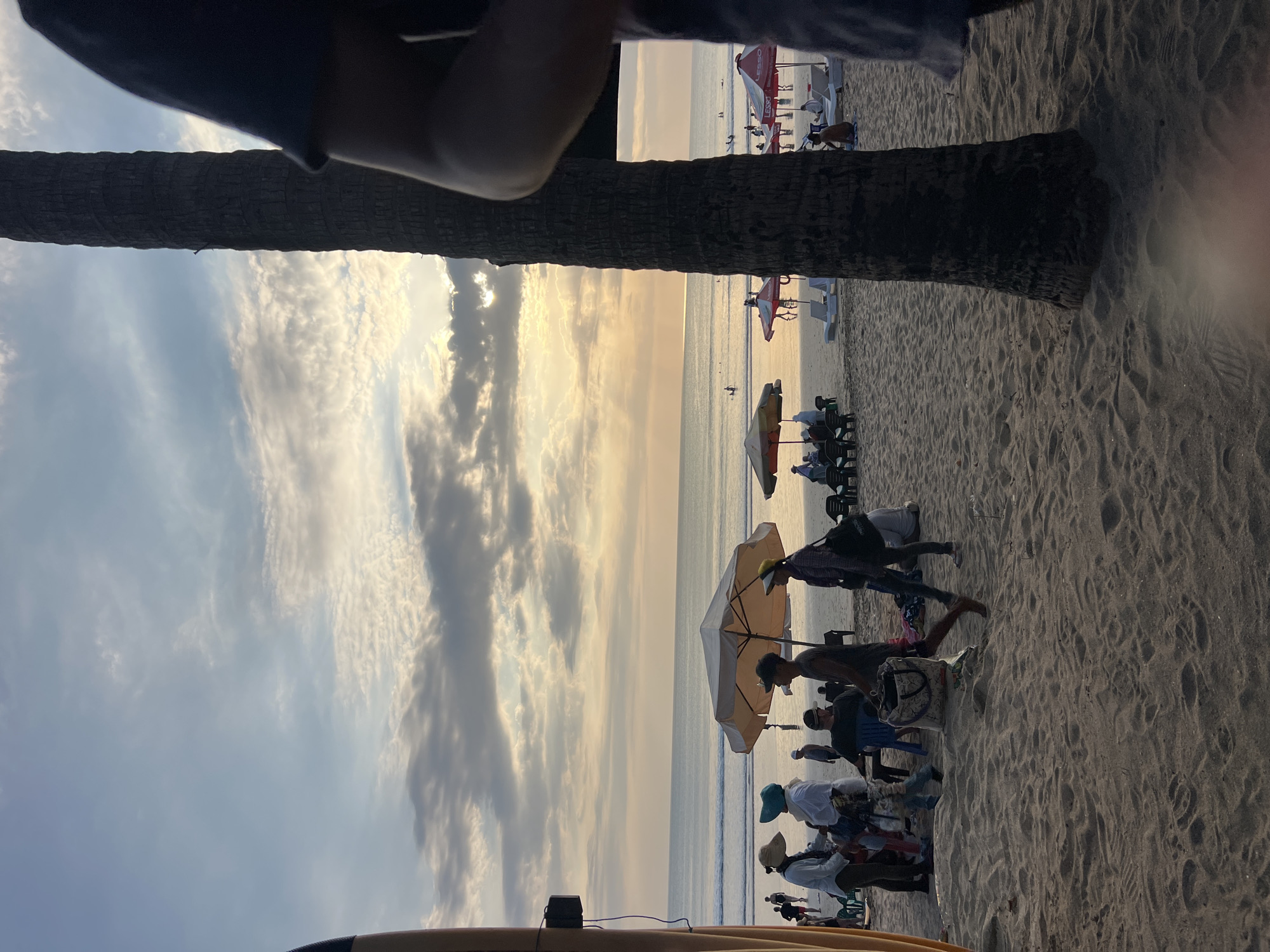 The image depicts a beach scene in Kuta, Indonesia, during sunset. The sky is filled with dramatic clouds and sun rays breaking through. The foreground shows sandy beach with footprints. Several people are visible on the beach, some walking, some sitting under umbrellas, and some gathered in small groups. There are beach umbrellas and lounge chairs set up. In the background, the ocean is calm with a few people swimming or surfing in the distance. A prominent palm tree trunk is in the right foreground, obscuring part of the view, and a surfboard is partially visible on the left. The overall atmosphere suggests a relaxed evening on a popular tourist beach.