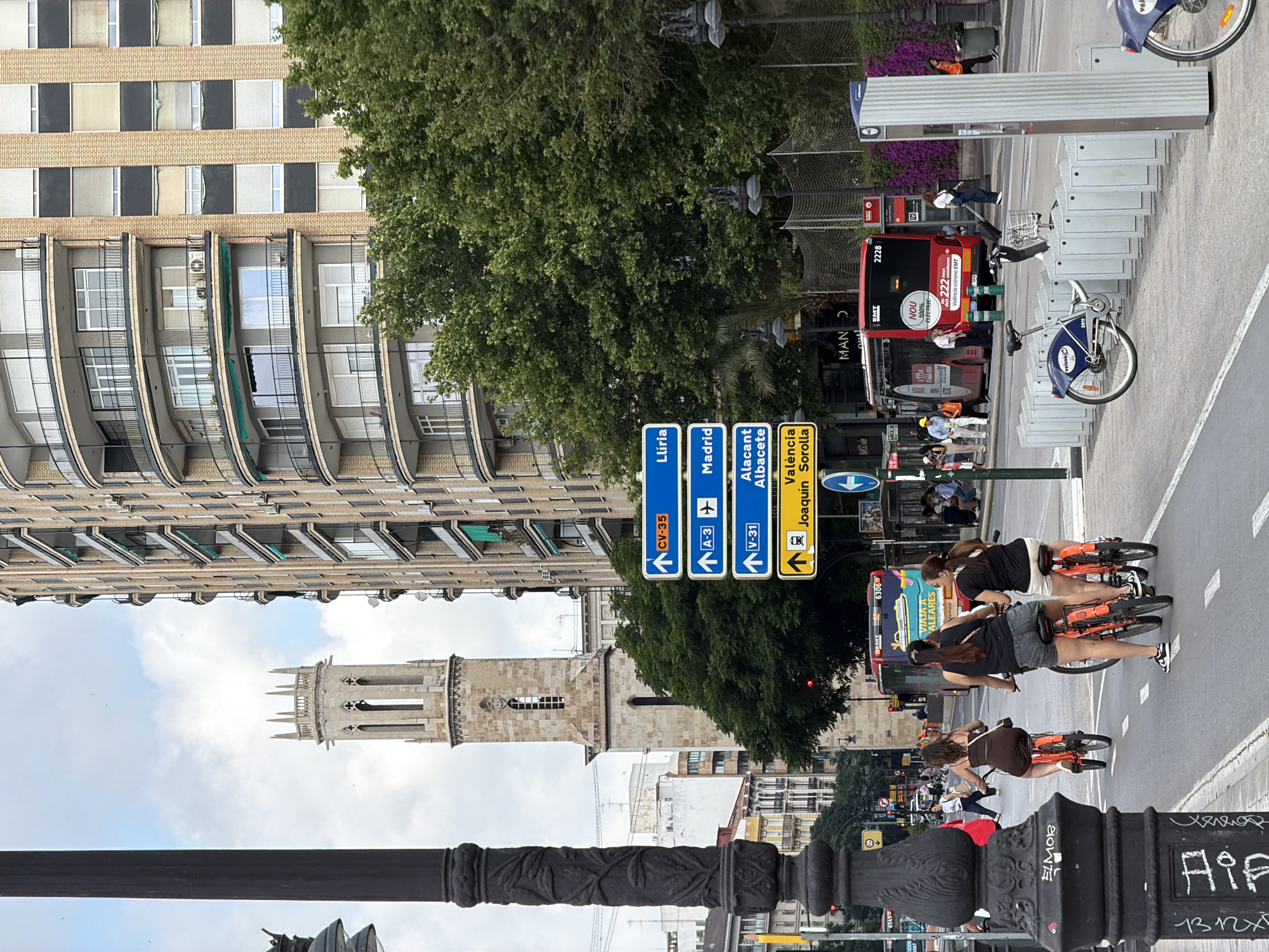 The image depicts a bustling urban street scene in Valencia, Spain, dominated by the bell tower of the Church of San Agustín in the background.
In the foreground, on the left, an ornate black lamppost with a detailed leafy design partially obscures the left side of the frame. Its base shows graffiti. To the right of the lamppost, three women are cycling orange bicycles in a designated bike lane. The lead cyclist wears a brown sleeveless top. The second cyclist wears a dark sleeveless top and denim shorts. The third cyclist wears a black sleeveless top and white shorts. All three are facing away from the camera, cycling forward.
Parallel to the bike lane, a red EMT (Empresa Municipal de Transports) city bus, numbered 2228, is visible. The bus features white text proclaiming "NOU 100% ELÈCTRIC" (New 100% Electric) and "València estrena EMT" (Valencia estrenates EMT). Several pedestrians are visible on the sidewalk to the right of the bus, and some are near its entrance.
Further to the right, a Valenbisi public bicycle rental station is present, with multiple blue bicycles docked. One blue Valenbisi bike, numbered 3917, is prominently docked.
Above the bus and Valenbisi station, a stack of four blue directional road signs is visible, mounted on a pole. From top to bottom, they indicate:
1. CV-35 Llíria (with an upward arrow)
2. A-3 Madrid (with an upward arrow and an airplane icon)
3. V-31 Alacant Albacete (with an upward arrow)
4. València Joaquín Sorolla (with an upward arrow and a train icon)
On the right side of the frame, a large, multi-story building with a curved facade and numerous balconies rises. The building features light brown brickwork and dark window frames, with some balconies having green awnings. A dense cluster of green trees obscures the lower levels of this building. Beyond these trees, a stone wall or fence and additional streetlights are visible.
In the background, against a partly cloudy, overcast sky, stands a tall, light-colored stone bell tower with a square base, gothic arched openings, and a decorative, crenellated top with smaller pinnacles. This is the bell tower of the Church of San Agustín. To the left of the tower, various urban buildings with diverse architectural styles are visible under the sky. The street is marked with white dashed lines for the bike lane.