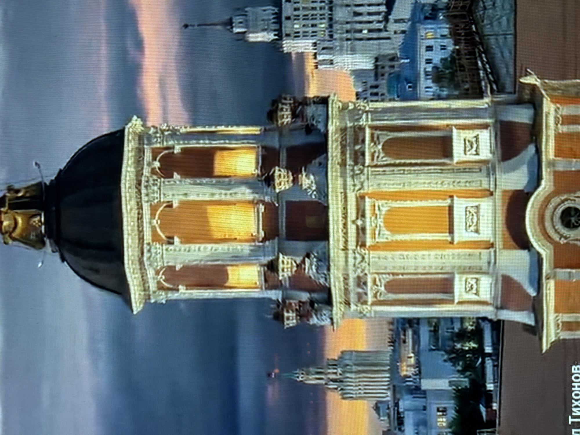 The image captures a close-up view of a decorative tower with a domed roof, likely a part of a church or historical building, against a backdrop of Moscow, Russia's skyline during twilight. The tower is illuminated from within, highlighting its ornate architectural details, including columns, arches, and intricate carvings. In the background, several tall buildings are visible, characteristic of Moscow's urban landscape, including what appears to be a Stalinist-era skyscraper with a spire. The sky is a gradient of deep blue and orange, suggesting the sun is either rising or setting. The foreground includes some text which is partially visible and illegible.