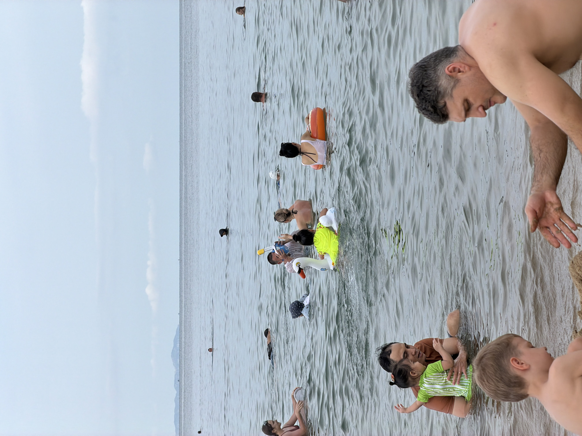 This image depicts a crowded beach scene in Nha Trang Ward, Vietnam, with numerous individuals engaged in water activities. The ocean is calm, with a hazy sky and distant landmass visible on the horizon. In the foreground, two children are playing in the shallow water, with one building a sandcastle. Nearby, a man is assisting a young girl in the water. Further out, several groups of people are swimming and wading, some with inflatable toys. A man in the middle ground is holding a snorkel and mask, and a child is sitting on a swan-shaped float. The overall atmosphere is one of leisure and recreation.