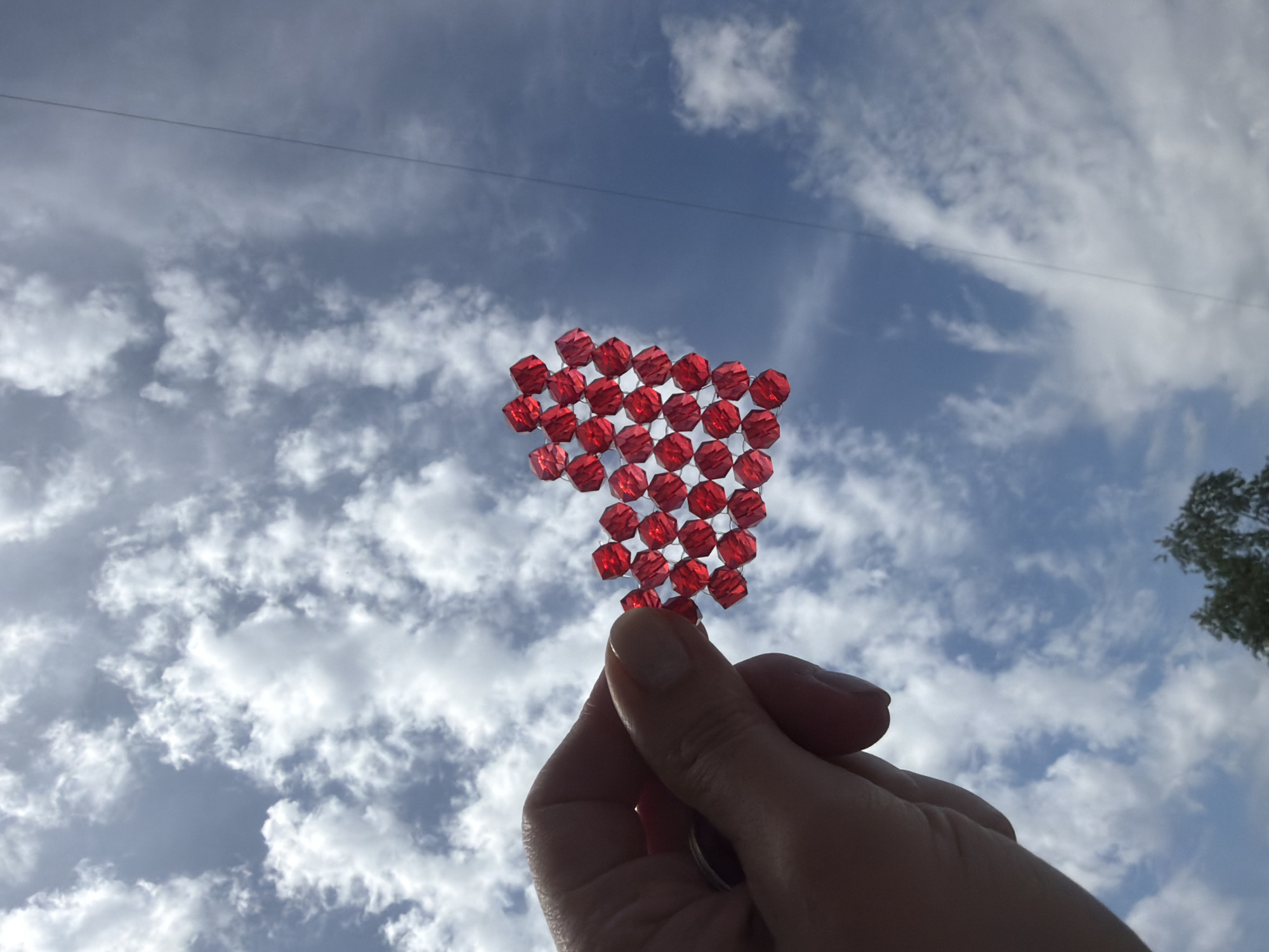 A hand holds a heart-shaped object made of red faceted beads against a background of blue sky with scattered white clouds. Part of a tree is visible in the upper right corner. A powerline extends horizontally across the top of the frame. The image lacks information to determine if the location is in Rostov-on-Don, Russia.