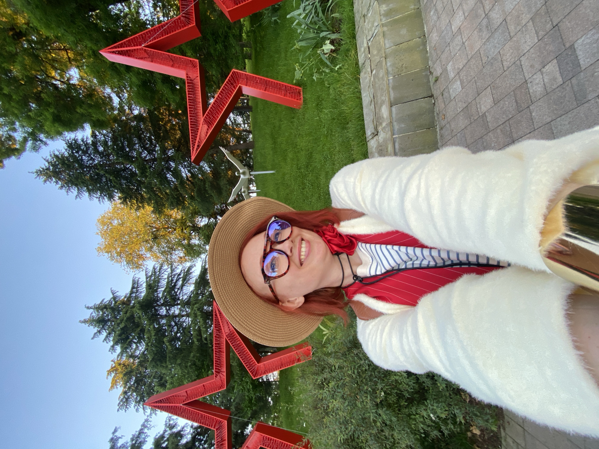 The image is a selfie featuring a smiling woman in an outdoor setting, likely a park or garden in Sochi, Russia. The woman, with reddish-brown hair, is wearing a straw hat, round framed glasses, a white fluffy cardigan or jacket, and a red and white striped top. She is looking directly at the camera with a broad, happy smile.
Behind her, prominent large red star-shaped structures are visible, suggesting a decorative installation or monument. These stars appear three-dimensional with an intricate, mesh-like texture. Further in the background, there is abundant green foliage, including tall evergreen trees and some deciduous trees displaying yellow leaves. A small, light-colored bird sculpture mounted on a pole can also be seen in the mid-ground. To the woman's right, a paved pathway made of gray rectangular tiles is partially visible.
The sky is clear and blue, and the warm lighting suggests the photograph was taken in the late afternoon or early evening. The overall scene conveys a pleasant and cheerful outdoor experience. No discernible text is visible on any of the objects.