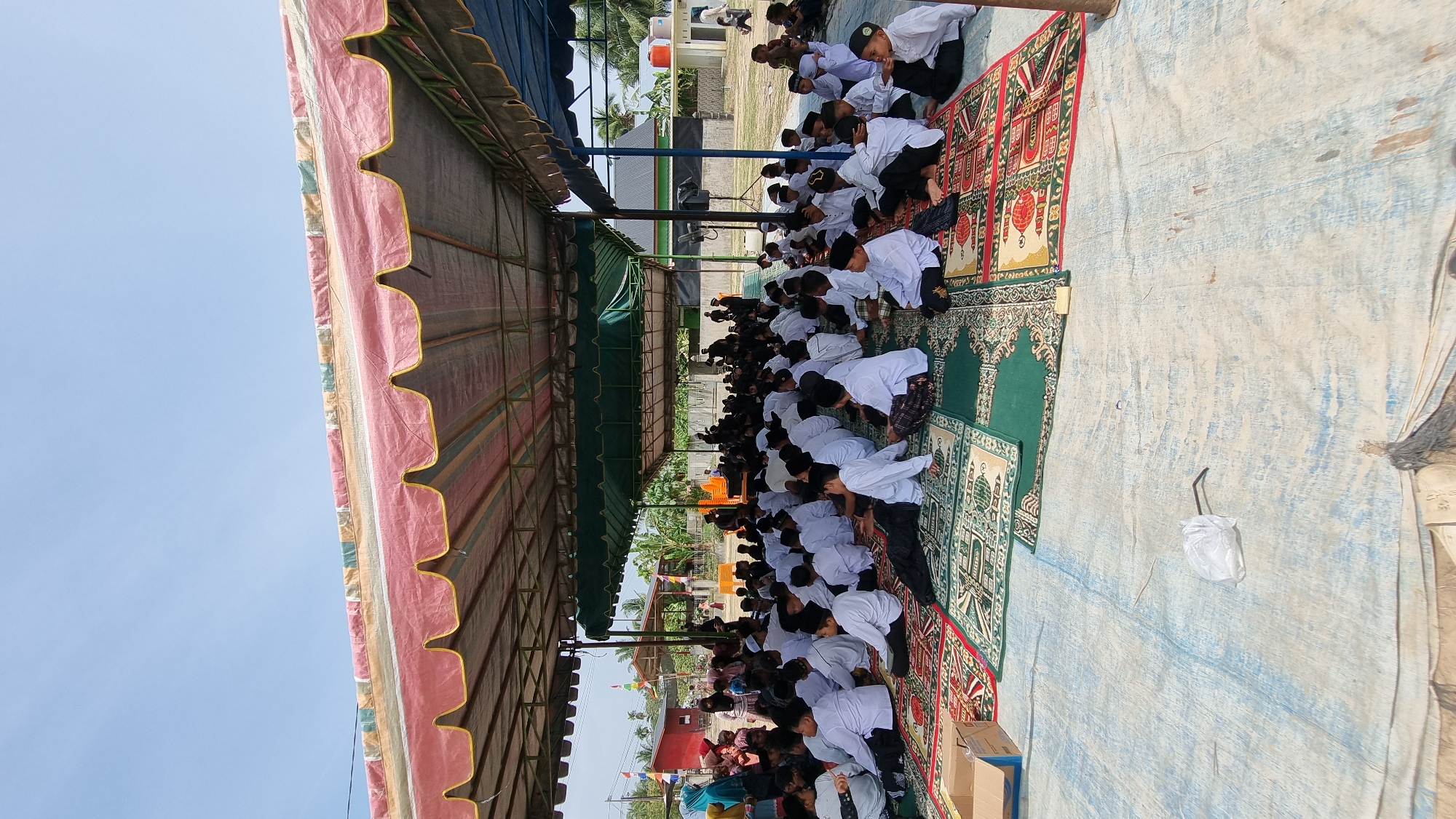 This image captures an outdoor gathering of many young boys, seated on colorful carpets beneath a large, striped tent. The boys are predominantly dressed in white shirts and black or patterned sarongs, with many wearing traditional peci caps. They appear to be listening attentively to something, possibly a lesson or religious observance. In the background, other adults and children are visible, as well as some buildings and trees, suggesting a village or community setting in Tumpokbeurandang, Indonesia. The ground beneath the tent is covered with a blue tarp, and there are various items scattered around, including a cardboard box and some white plastic bags. The scene is illuminated by bright sunlight, indicating it is daytime.