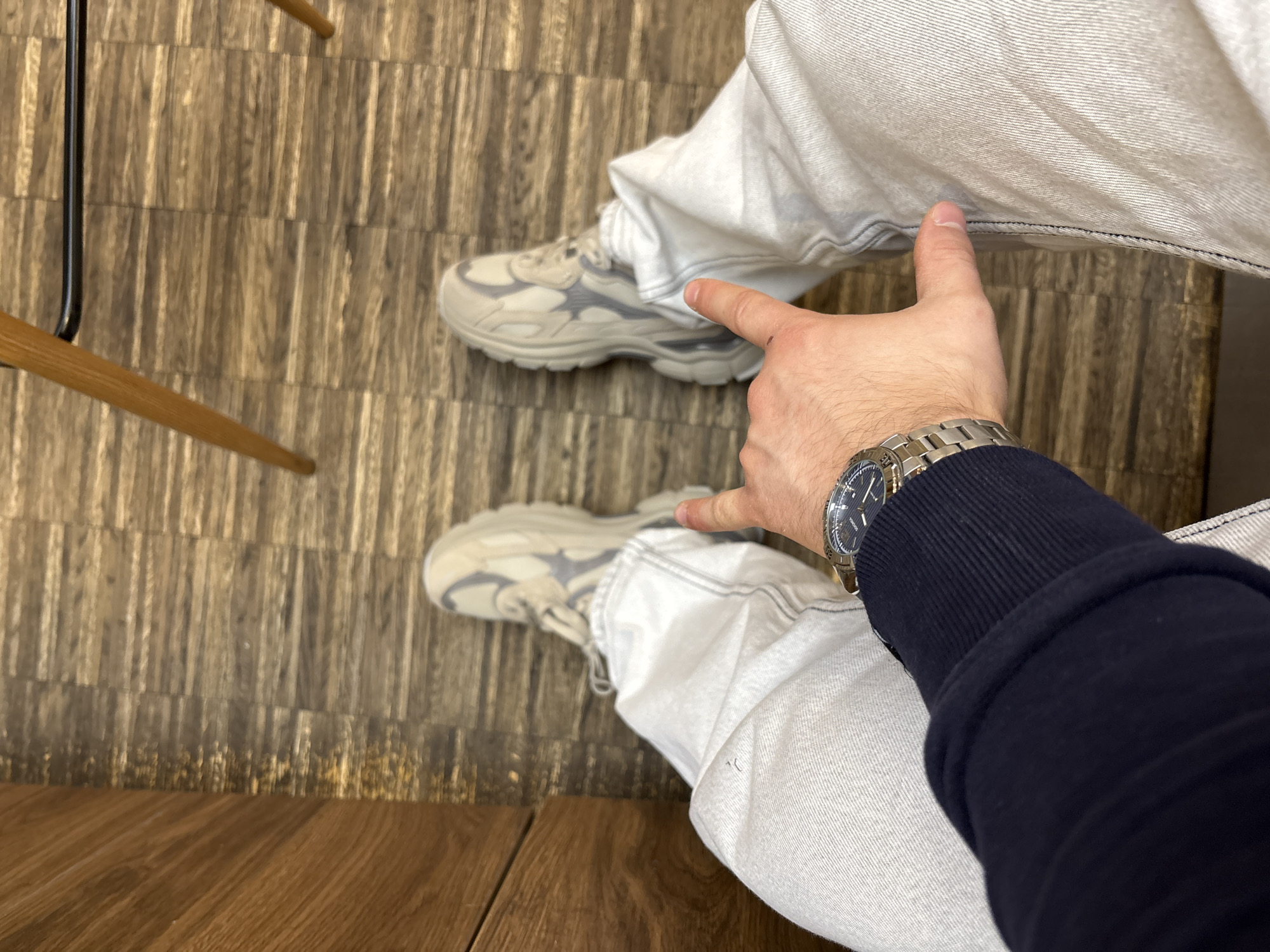 A high-angle shot reveals the lower body of an individual clad in light-wash denim jeans, adorned with a silver watch on their left wrist, and a navy blue sleeve. The person is wearing gray sneakers. Their left hand is extended, the index finger pointing towards the right knee. They are sitting on a chair. The background displays a wooden floor of light and dark brown color. The location is Sofia, Bulgaria.