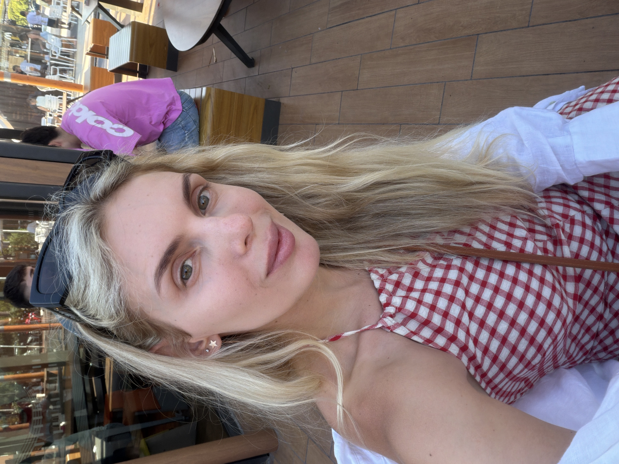 A woman is taking a selfie outdoors, likely in a casual dining area. She has long blonde hair, is wearing sunglasses on top of her head, and has star-shaped earrings. Her top is red and white gingham. Behind her, a person in a pink shirt is visible, and further in the background, there are tables and chairs, suggesting a restaurant or cafe setting. The floor is made of wooden tiles. The lighting indicates it is daytime. The location is described as Aldaia, Spain.