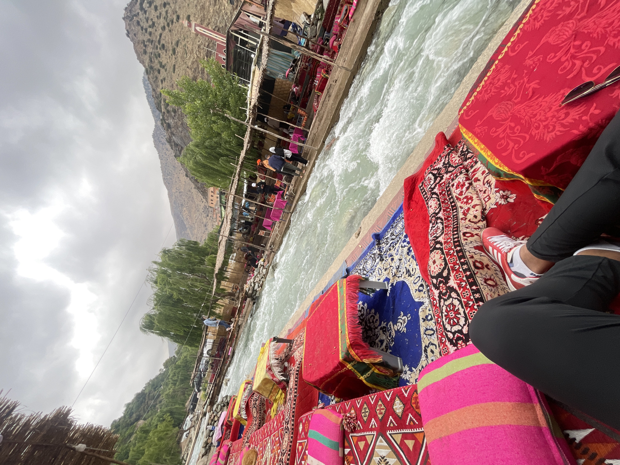 A first-person perspective photo shows a stream of water coursing through what appears to be a bustling area in Asgaour, Morocco. The foreground is occupied by colorful cushions and rugs, with a person wearing black pants and red and white sneakers sitting among them. Sunglasses lie on a red patterned surface. The stream is bordered by structures adorned with colorful textiles and people are visible walking along the structures beside the stream. Lush greenery covers the hillside in the background, which leads up to rocky terrain and a cloudy sky.