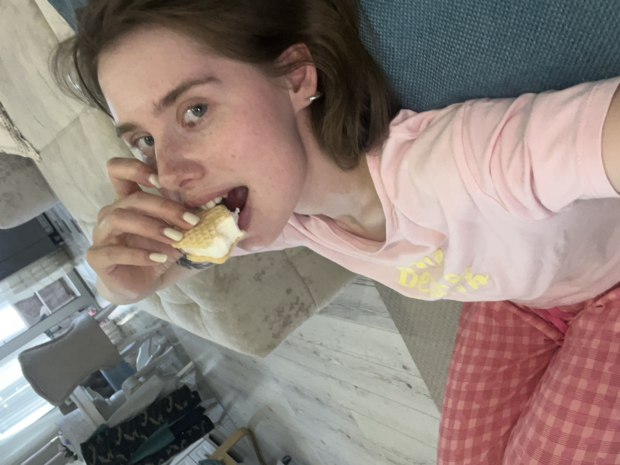 A young woman is lying on a couch, taking a selfie while eating an ice cream sandwich. She is wearing a pink t-shirt and pink checkered pajama pants. Her hair is brown and short. The couch is blue. The background includes a window and other furniture. The location is likely indoors, but no specific details confirm it is Chaikovsky, Russia.