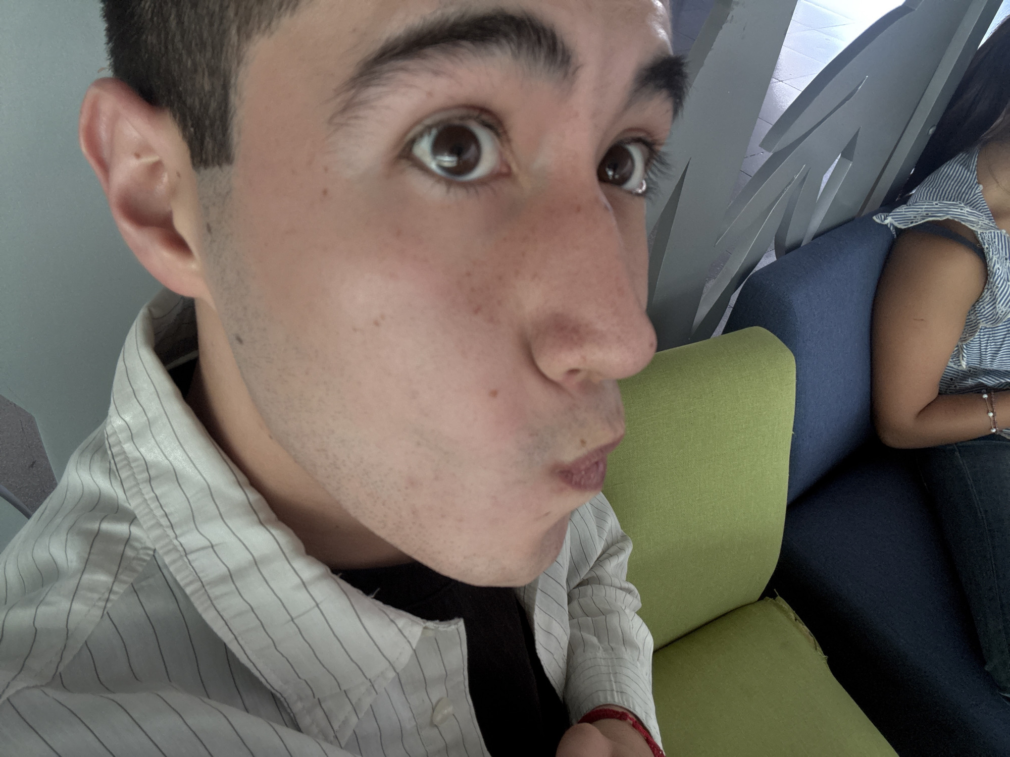 A close-up, selfie-style photograph shows a young man with a light complexion, dark hair, and brown eyes looking slightly off-camera with a puckered expression. He is wearing a black t-shirt under a white and light gray striped button-down shirt. The background includes a person sitting on a blue chair and a green cushioned object, possibly part of the seating arrangement. In the upper right corner, a design feature consisting of gray vertical elements is visible. The location is suspected to be Puebla, Mexico, based on visual clues not explicitly present in the photograph.