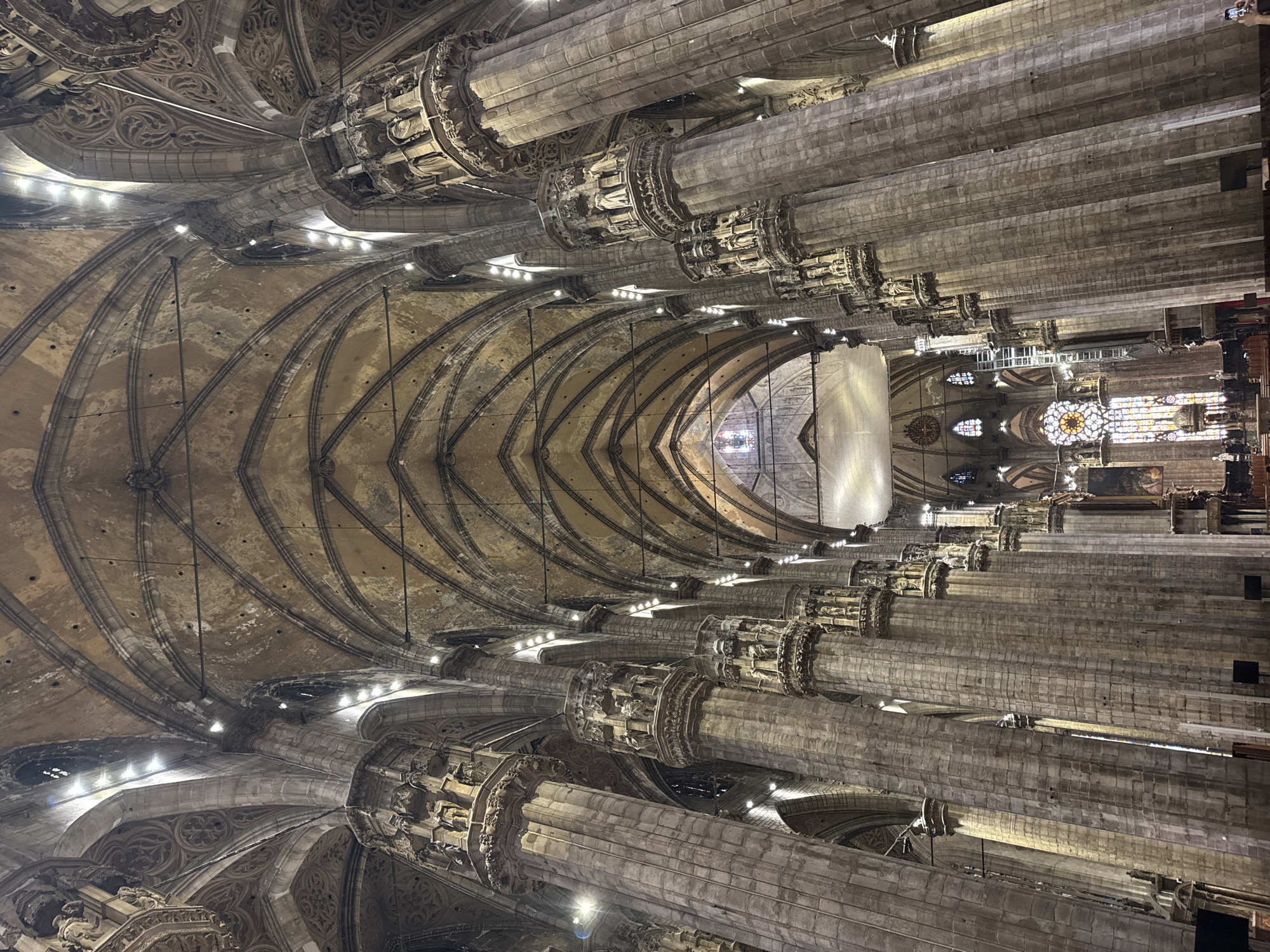 The image shows the interior of a grand cathedral, likely the Duomo di Milano, in Milan, Italy. The perspective is from the nave, looking towards the apse. Massive, intricately carved stone columns with Gothic-style capitals rise to support a vaulted ceiling with ribbed arches. Natural and artificial lights illuminate the space, highlighting the aged stone and ornate details. In the distance, stained-glass windows and what appears to be an altar or chancel area are visible. The scene is quiet and appears to be during daylight hours, with no visible people in the immediate foreground or mid-ground, though the presence of lighting suggests it may be a public space.