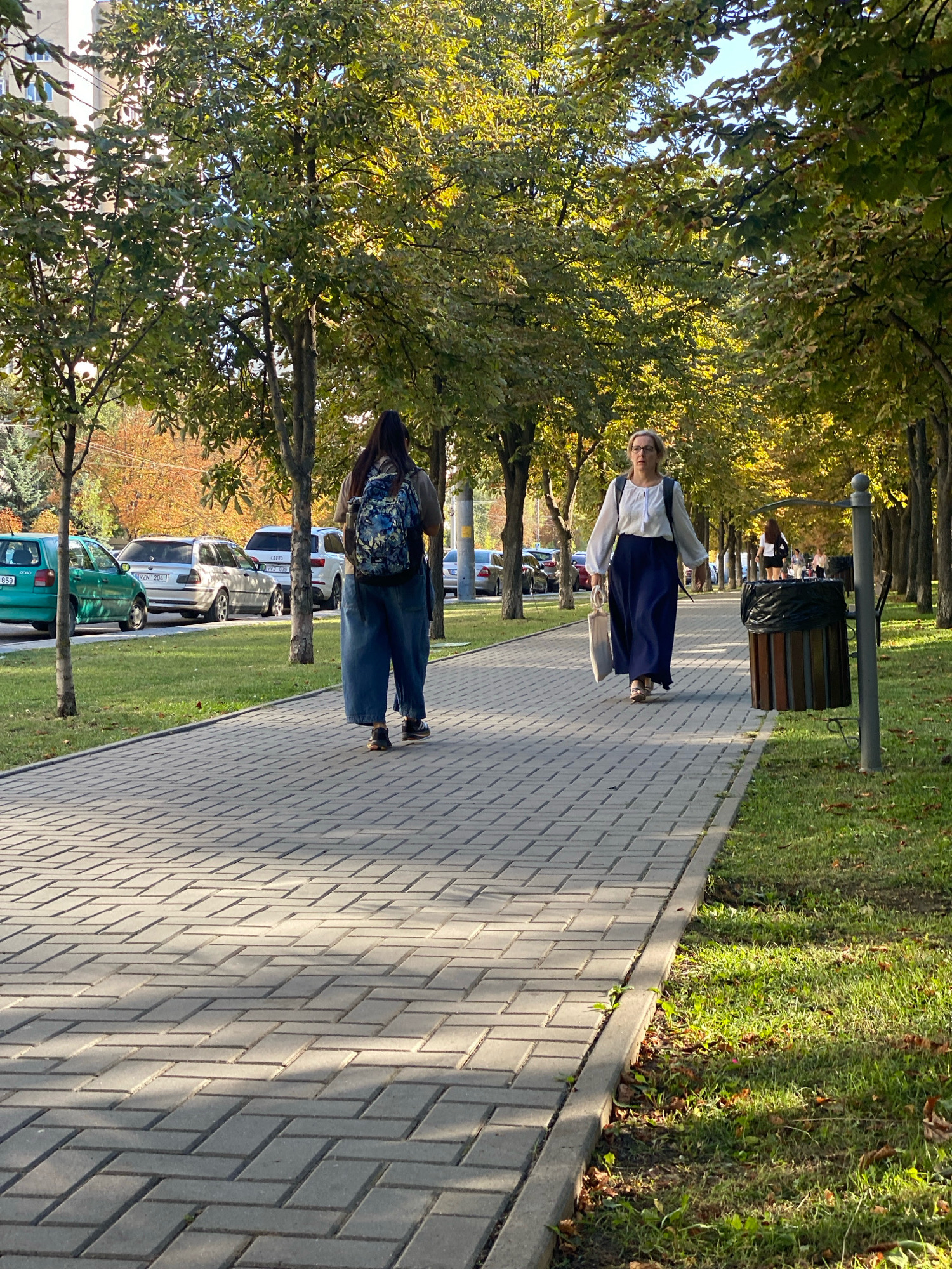 The image shows a sunny day in a city park or along a tree-lined boulevard. Two women are walking on a paved sidewalk. The woman in the foreground, on the right, is wearing a white blouse and a long blue skirt and carrying a light-colored bag. The woman in the background, on the left, is wearing a backpack and wide-legged pants. Cars are parked along the street on the left side of the image, and trees with autumn foliage line the right side. A trash can is visible near the right edge of the image. The overall scene suggests a pleasant, everyday urban environment, likely during the fall season. There is no direct evidence within the image to confirm the location as Chișinău, Moldova.