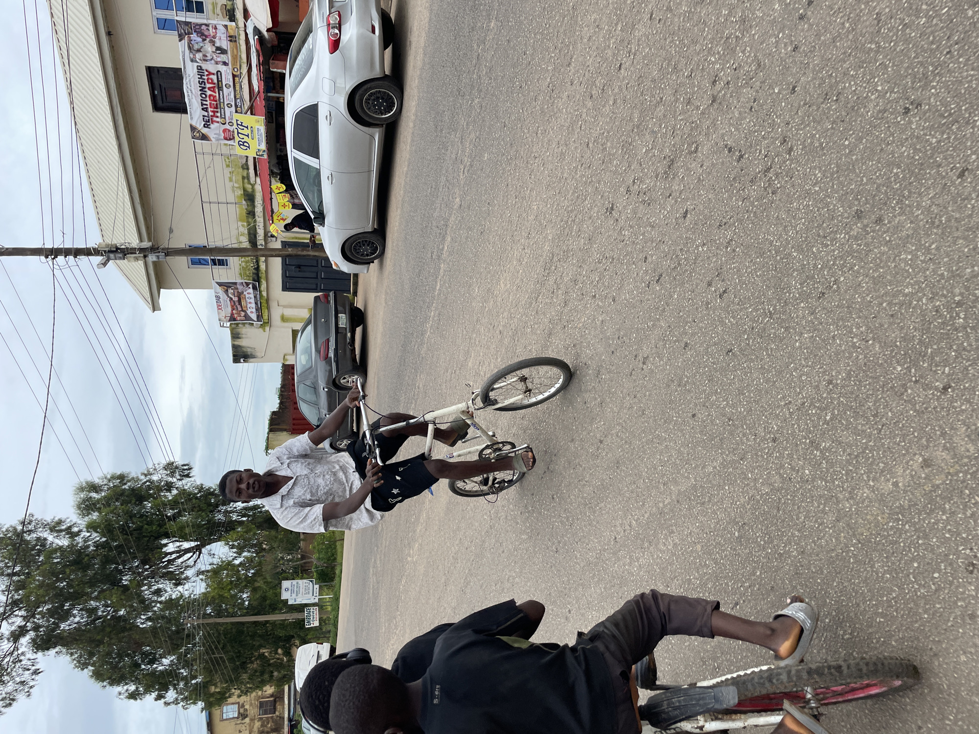 The image depicts a street scene in Jos, Nigeria, featuring a young man riding a small bicycle in the foreground. He is wearing a white patterned short-sleeved shirt and black shorts with white star patterns. He has on a pair of silver slide sandals. Behind him, another young person is seated on the back of a bicycle, wearing a black t-shirt and dark trousers. Two bicycles are partially visible in the foreground, one being ridden by the young man and the other carrying the second individual.
The street is paved and appears to be a typical urban road. To the right of the road, there is a two-story building with a pale cream facade. Banners are displayed on the building; one prominently reads "RELATIONSHIP THERAPY" with additional text below it. Another banner to the left of this one has the text "JOEDAB" and images of people. Further down the street, parked cars are visible, including a silver sedan on the right and a dark sedan to the left, in front of a building with dark doors. In the background, tall trees with green foliage rise against a cloudy sky. Utility poles with wires are also present along the roadside. The overall impression is of a busy street in a Nigerian city.
