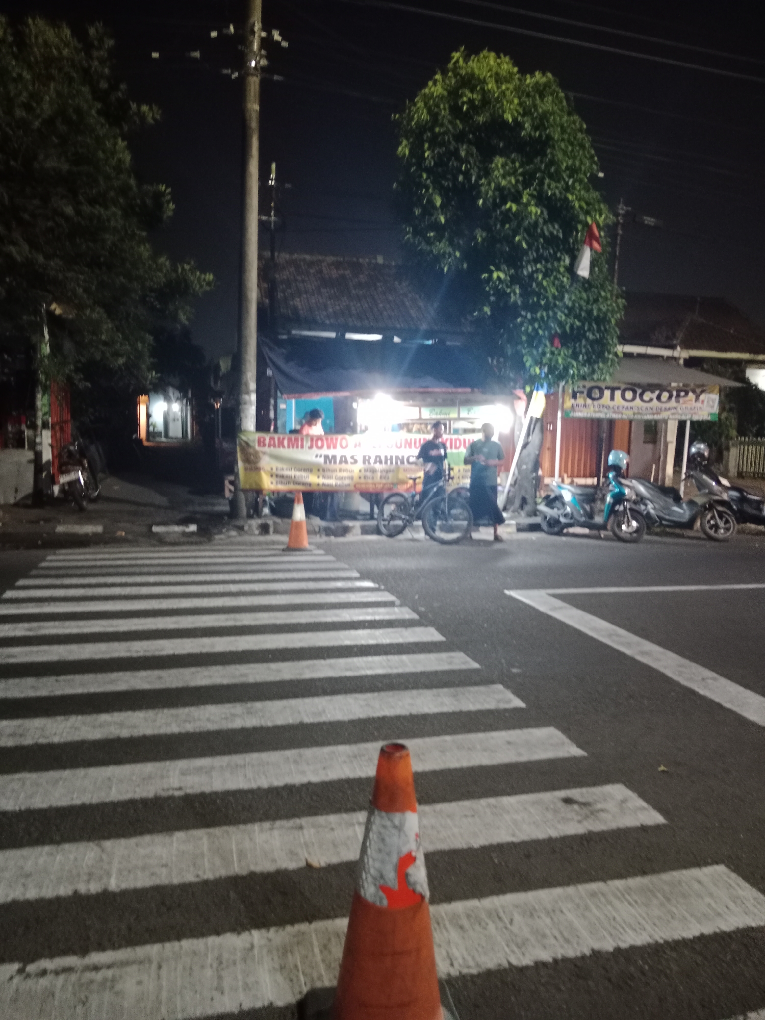 A night-time scene depicts a busy street in Yogyakarta, Indonesia, with a pedestrian crossing in the foreground. A traffic cone, orange and white, stands on the right side of the pedestrian crossing. In the background, several people are gathered around food stalls. One stall has a banner that reads "BAKMI JOWO" and "MAS RAHMO". Another stall has a banner that reads "FOTOCOPY". Several motorcycles are parked on the side of the road. The lighting is dim, suggesting it is late at night. A large tree with green leaves is visible on the left side of the image. The overall impression is that of a lively street scene in a city that never sleeps.