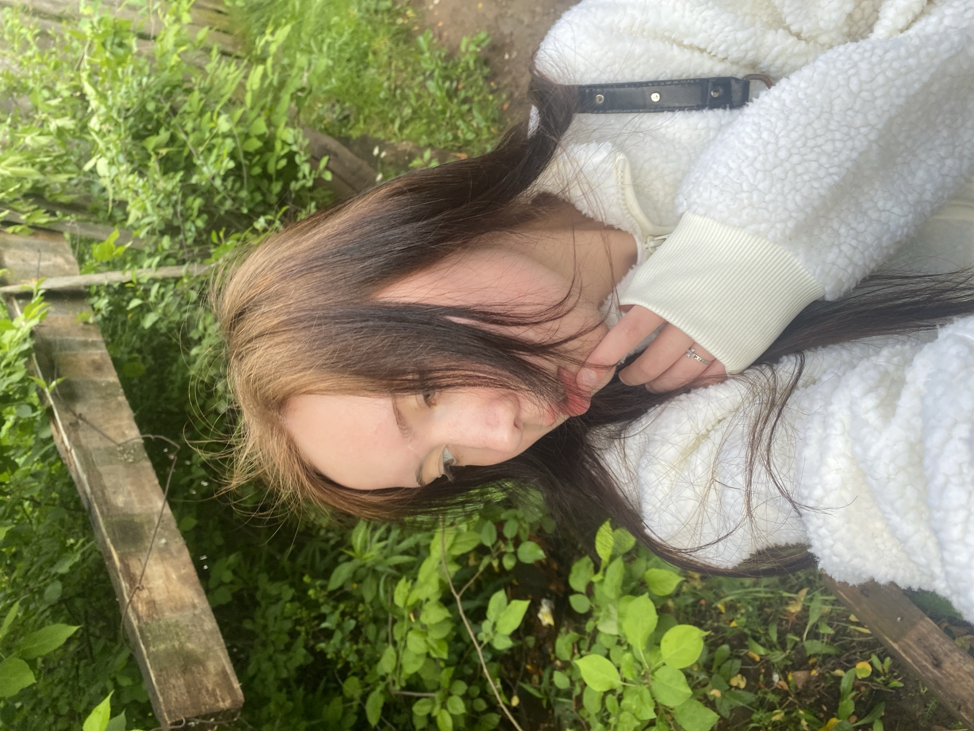 A person is partially seen in an outdoor setting near greenery and a wooden structure. The individual has long, brown hair with lighter strands at the top, and is wearing a white, fluffy jacket with a ribbed cuff at the wrist. Their left hand is visible, adorned with a ring. The scene is dominated by lush green vegetation surrounding what appears to be a weathered wooden plank or fence. The location seems to be outdoors with natural elements, but the specific location in Unknown, Unknown cannot be determined from the given image.