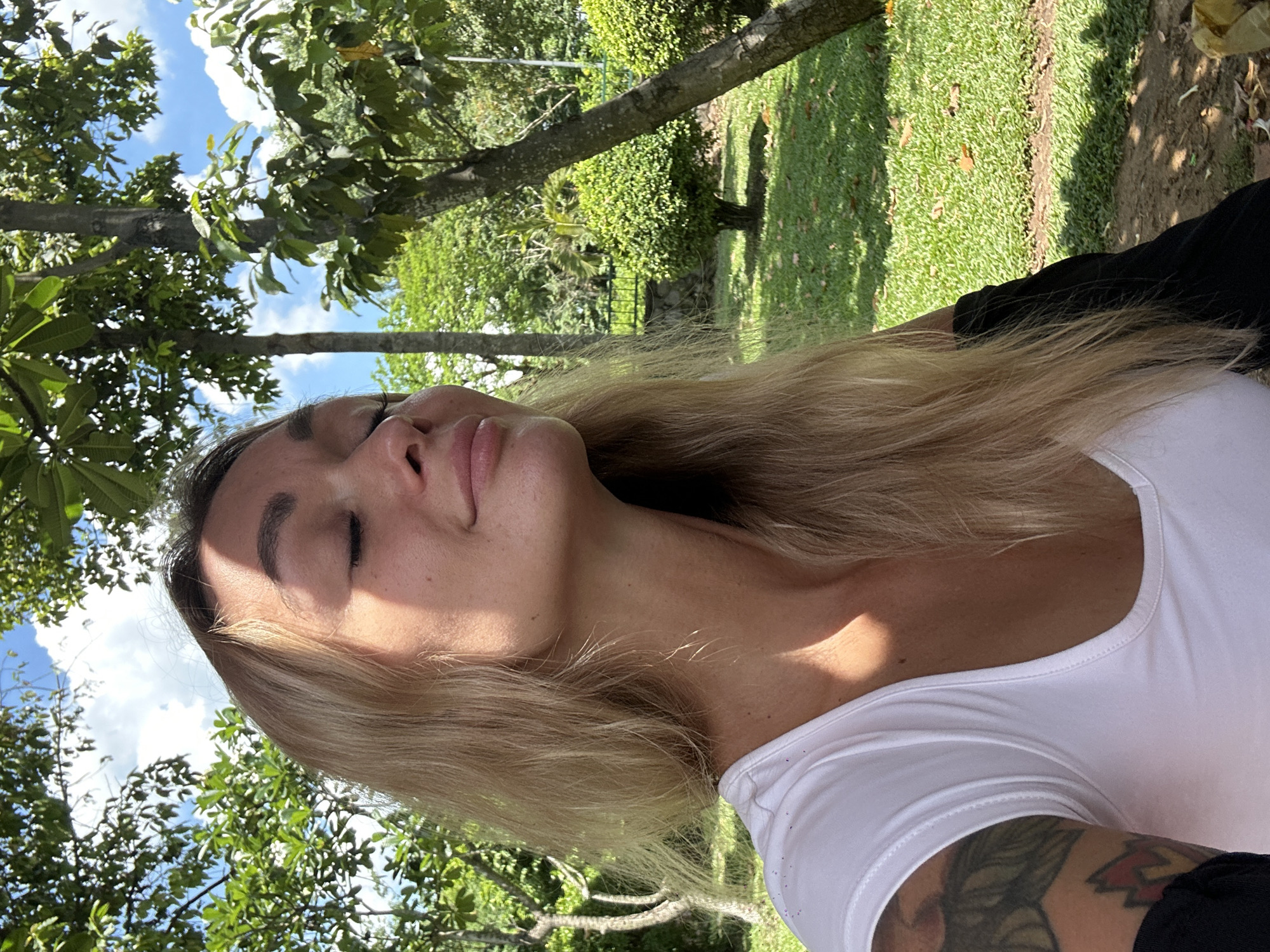 A woman with light blonde hair and closed eyes is outdoors. She wears a white shirt and a black garment visible on the right. A tattoo is partially visible on her right arm. The background shows trees, grass, and a glimpse of a blue sky with clouds. The location could be a park or garden in Dĩ An, Vietnam.