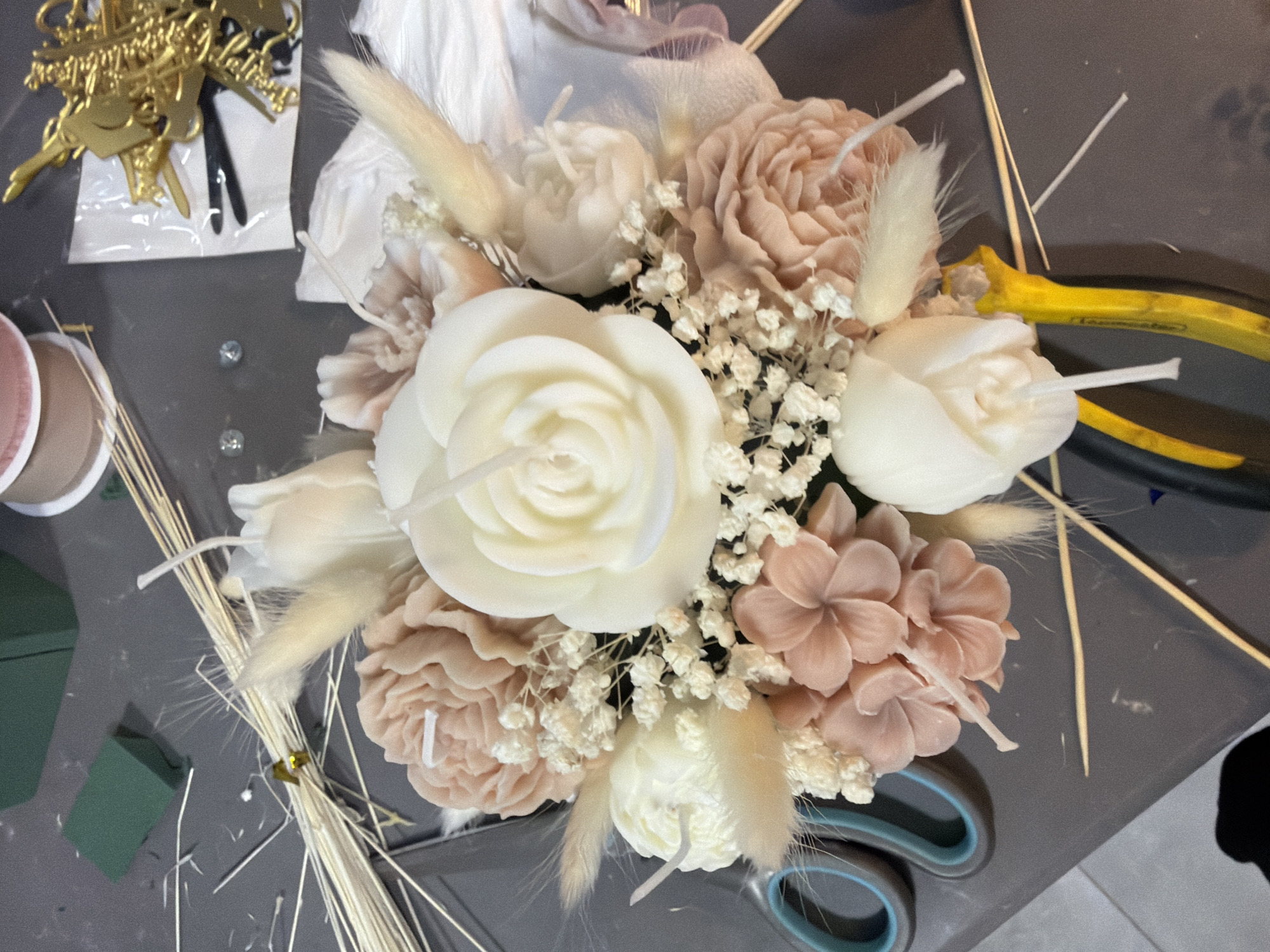 This is a close-up, high-angle shot of a flower arrangement, possibly in the process of being made, likely indoors. The arrangement features various types of flowers, including roses and other blooms in shades of white, cream, and light peach. Some of the flowers appear to be candles, as evidenced by the visible wicks. There are also sprigs of baby's breath and delicate, feathery pieces interspersed among the larger flowers.
Several objects are scattered around the floral arrangement on a grey surface. To the upper left, there's a gold "Happy Birthday" cake topper in its packaging, along with a spool of ribbon. Bundles of slender twigs are visible on the left side. To the right, a pair of yellow-handled pliers and scissors with teal handles are visible. Green floral foam is visible in the lower left.
There's no evidence that this image was taken in the city of Novo Selo, Bulgaria.