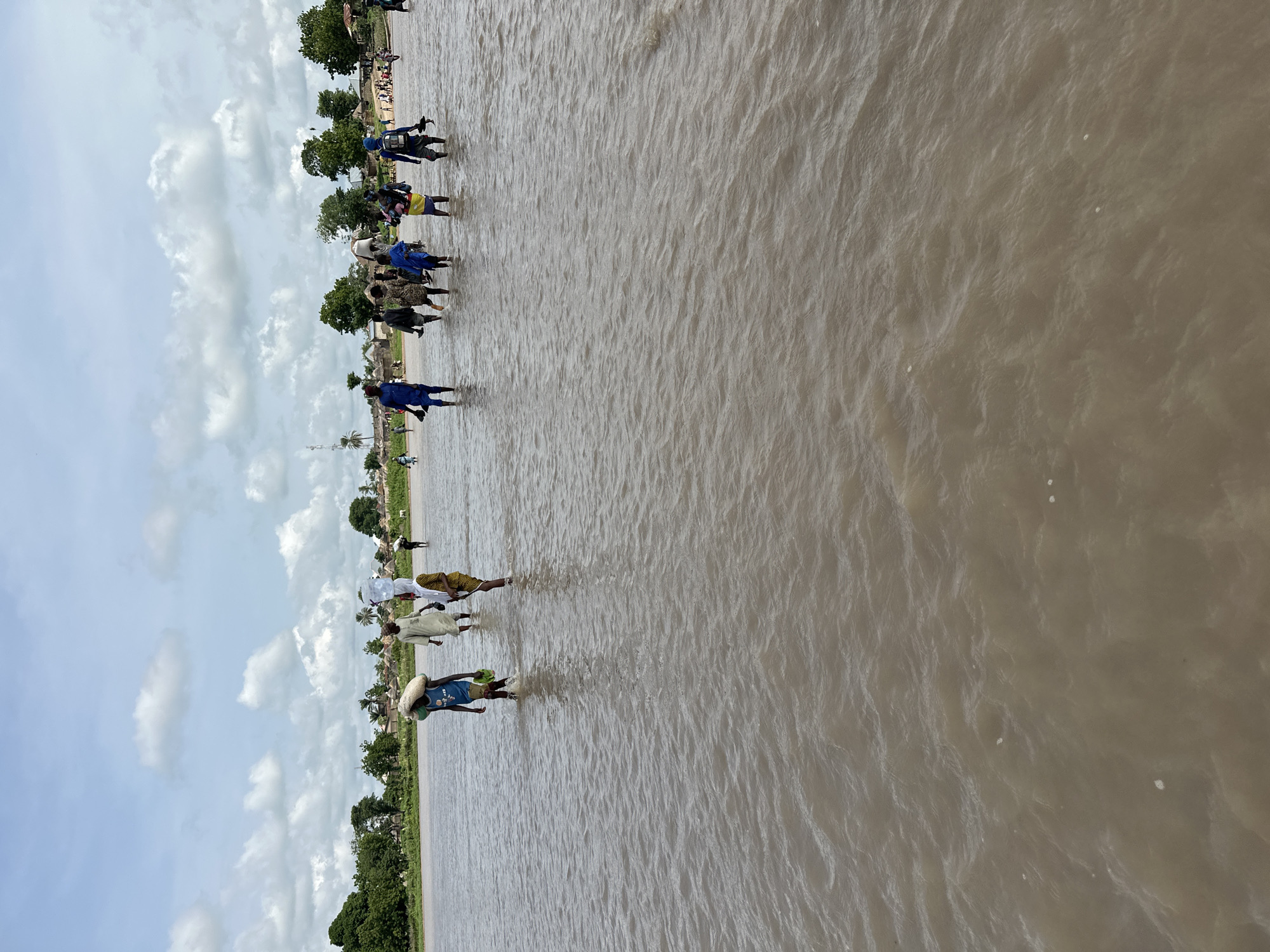 A group of people are wading through a wide, shallow river under a cloudy sky. Many of them are carrying items on their heads, including what appear to be large containers and sacks. The riverbanks are lined with trees and small buildings, suggesting a rural or semi-rural setting, likely in Kunini, Nigeria, given the context. The water is murky brown and appears to be flowing gently. The people are dressed in casual clothing, with some wearing traditional attire. The overall scene depicts a daily activity of crossing the river, likely for transportation or to access resources.