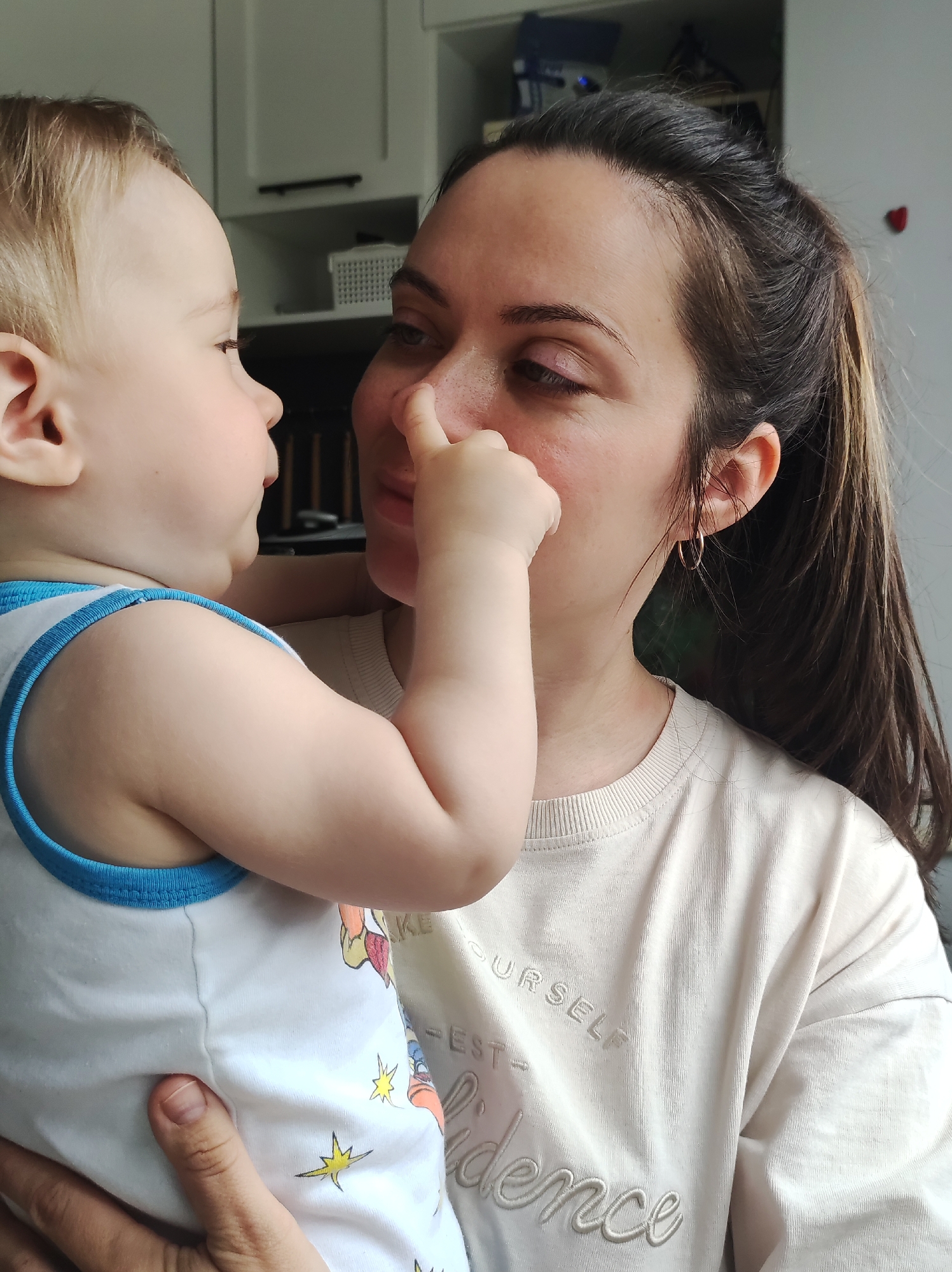 A medium shot shows a woman holding a young child. The child, facing left, touches the woman's nose with its right index finger. The woman, facing right, looks down at the child. She has brown hair pulled back in a ponytail, hoop earrings, and is wearing a beige shirt with embossed writing. The child wears a white tank top with blue trim and cartoon graphics. The background features white cabinets. The location is Chaikovsky, Russia.
