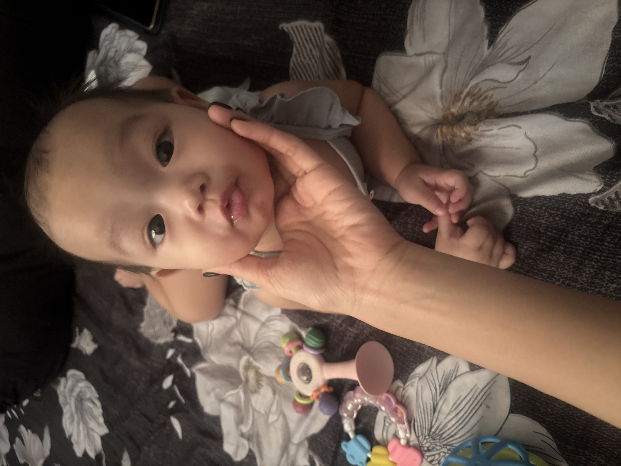 An overhead shot captures an infant lying on a floral-patterned bed. The infant's face is the primary focus, with dark eyes, a small nose, and slightly parted lips. A person's hand gently supports the infant's chin and cheek. In the foreground, scattered on the bed, are several baby toys, including a pink rattle with colorful attachments and a bracelet-like toy with brightly colored animal shapes. The background is softly blurred, with a dark, possibly upholstered surface visible. The scene is indoors, likely in a residential setting. The specific location in West Jakarta, Indonesia, cannot be determined from the visual information provided.