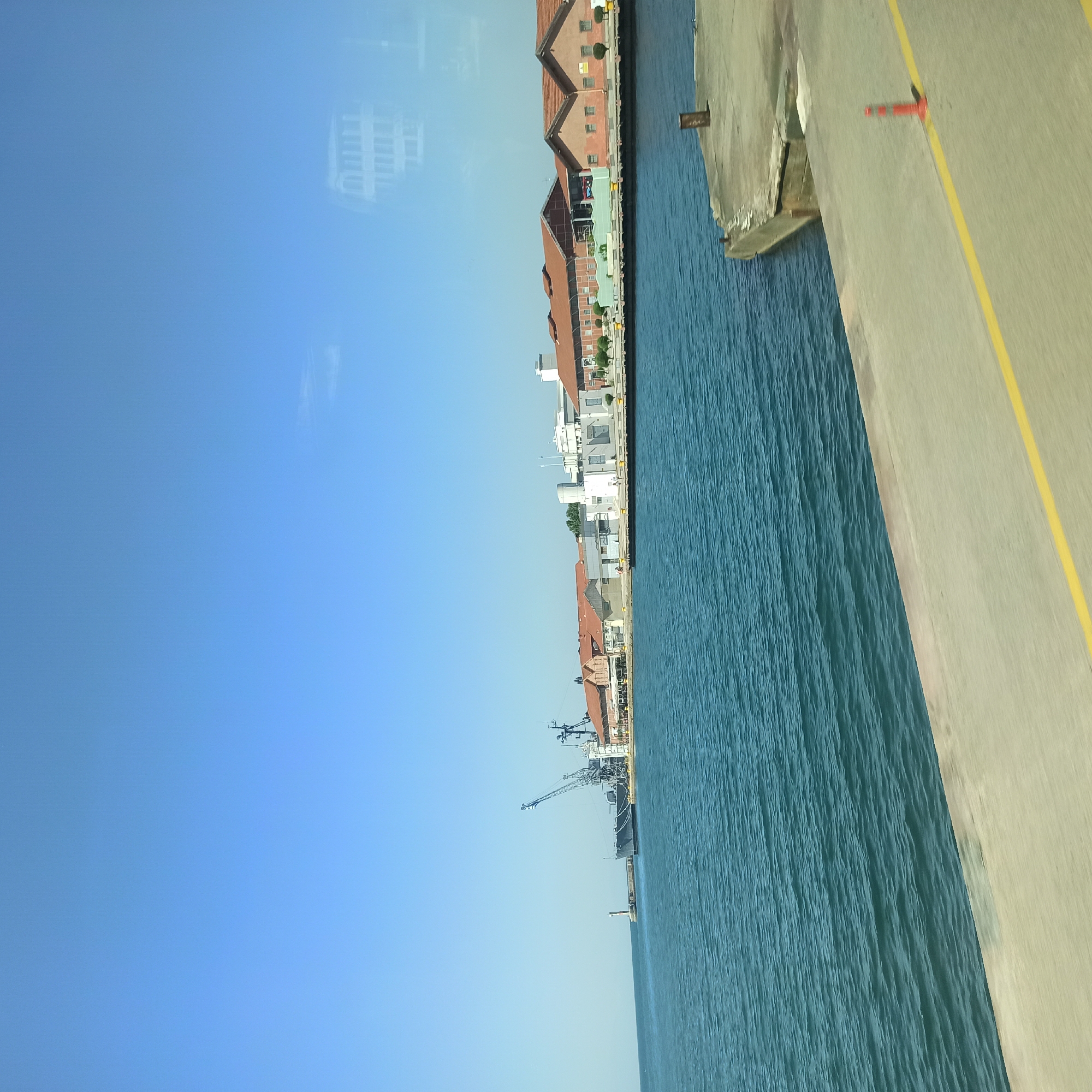 The image depicts a view of the port area in Thessaloniki, Greece. In the foreground, a concrete quay runs diagonally across the frame, marked with a yellow line and an orange traffic cone. The quay is adjacent to the blue water of the harbor, which exhibits gentle ripples. In the middle ground, a line of buildings with red-tiled roofs and light-colored facades stretches along the waterfront. These buildings appear to be industrial or commercial structures. A large ship, possibly a naval vessel with a crane, is docked near a lighthouse situated on a pier in the distance. The sky above is clear and bright blue. The overall scene suggests a working port under clear weather conditions.