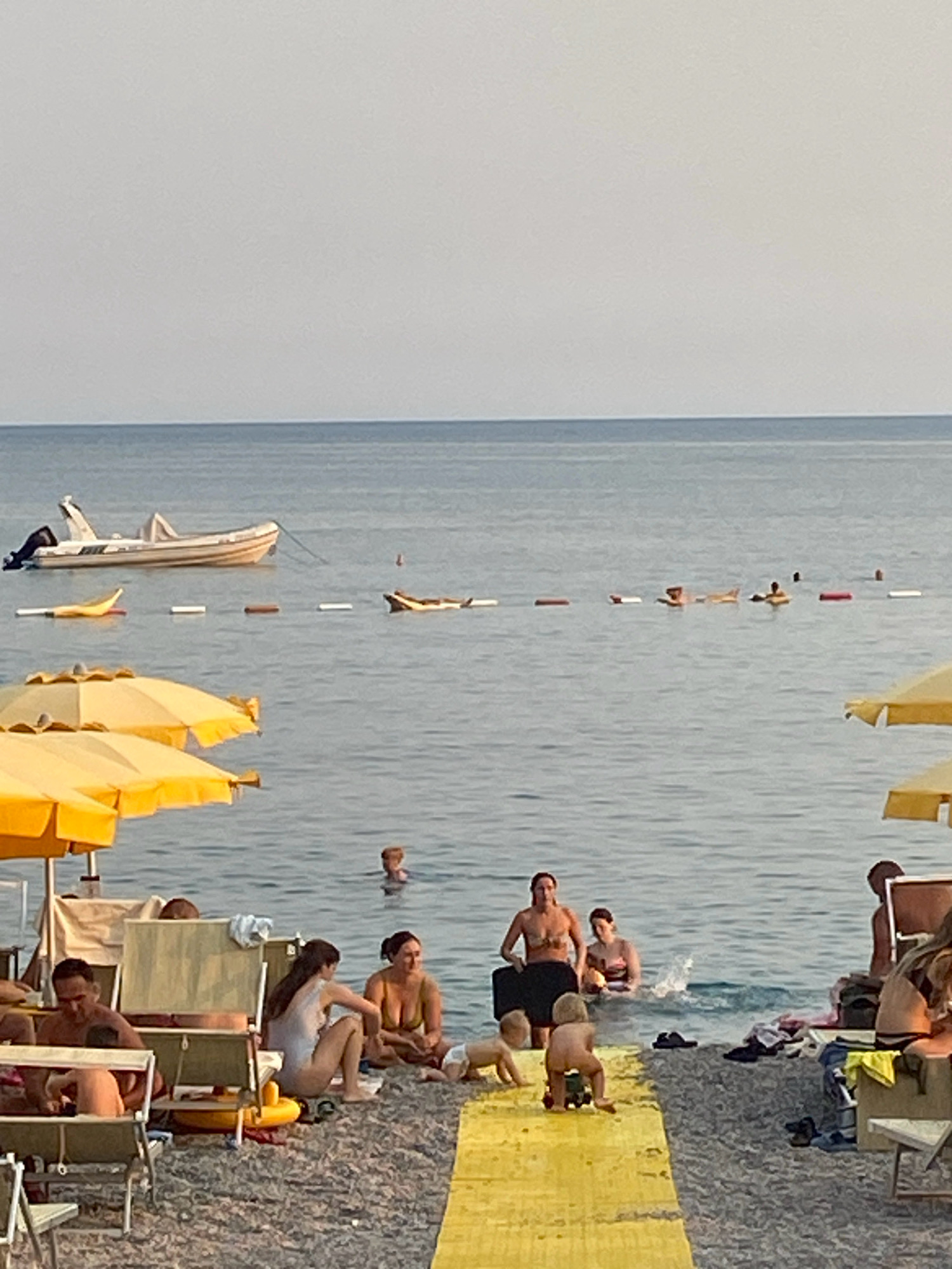 The image captures a lively beach scene in Prijevor, Montenegro, on a hazy, overcast day. The calm sea stretches out towards the horizon, dotted with a few small boats and inflatable toys. On the shore, beachgoers relax under yellow umbrellas, with some wading in the water and others sitting on the pebble beach. Children play near the water's edge, and a couple of adults appear to be sunbathing or resting on lounge chairs. The overall atmosphere is one of leisure and relaxation, with a hint of a vacation ambiance.