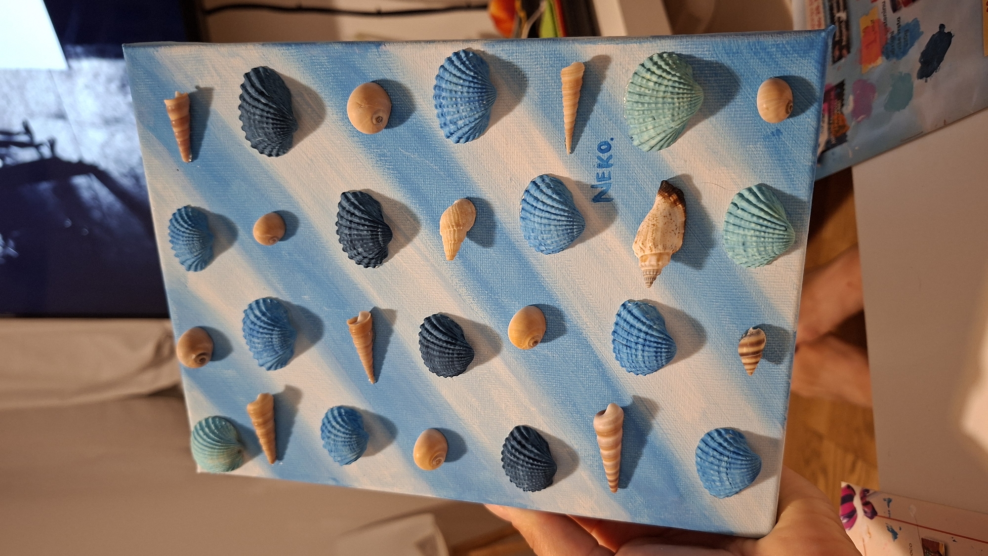 The image shows a close-up of a piece of art made with seashells glued onto a canvas painted with blue and white diagonal stripes. The canvas is held by a person's hand. There are multiple seashells of various shapes, sizes, and colors, including blue, teal, orange, and tan. Some shells are round, while others are elongated or fan-shaped. A word, "NEKO," is written in blue on the canvas. The background is blurry, showing a room with a television screen in the upper right corner and a curtain on the left. The lighting suggests an indoor environment. There is no visible indication of the location being in the city of Podgorica, Montenegro.