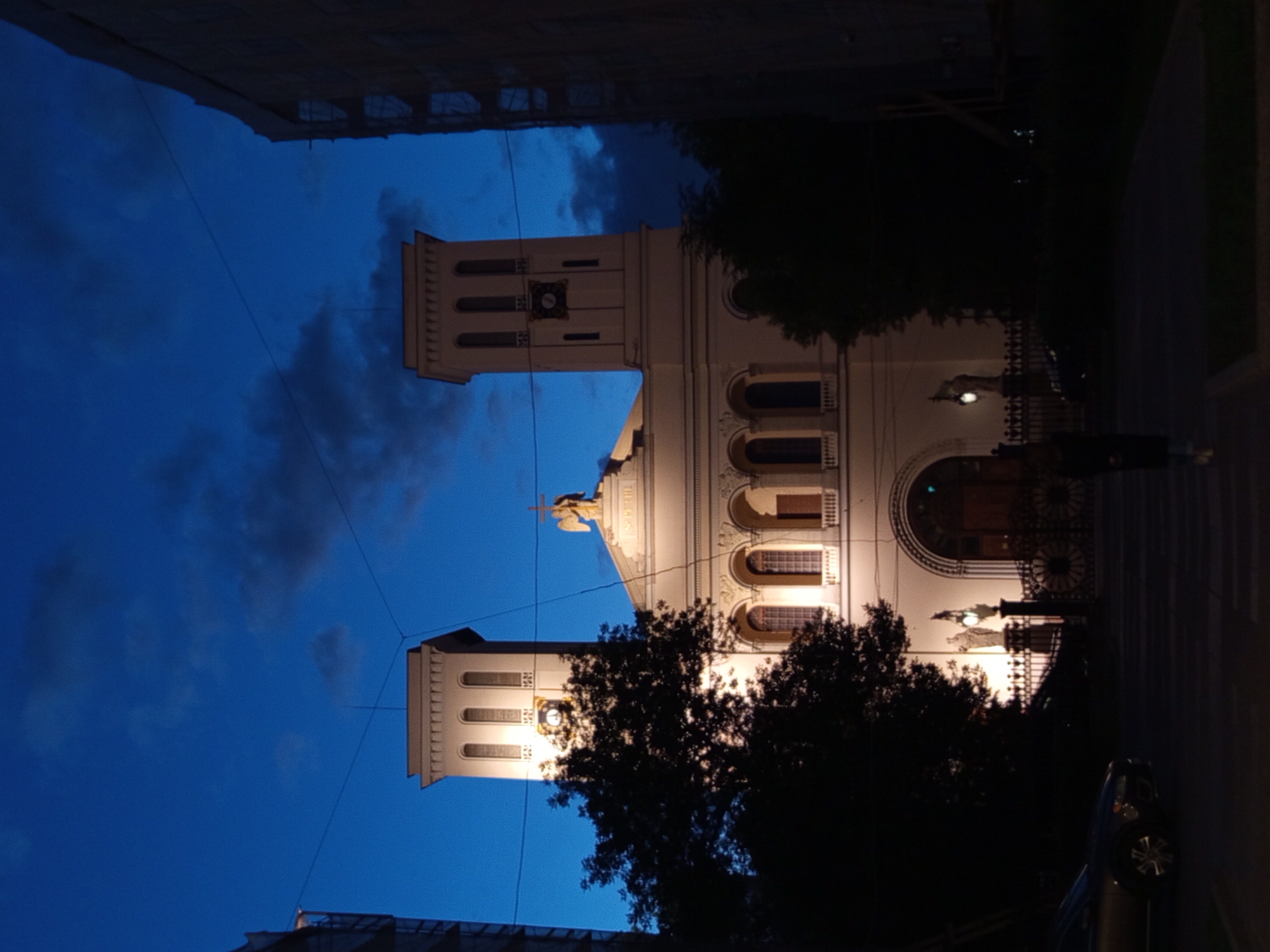 A low-angle, vertical, outdoor shot captures the facade of a building, likely a church or a civic building, in Saint Petersburg, Russia, during twilight. The sky is a deep, bruised blue with scattered dark clouds. The building is illuminated by artificial lights, highlighting its cream-colored stone and architectural details. It features two prominent square towers with arched windows and a clock on the left tower. A smaller arched structure sits between the towers, topped with a statue of an angel holding a cross.
In the foreground, a dark-leafed tree partially obscures the left side of the building. To the far left and right, the edges of taller, modern buildings frame the scene. A dark-colored car is parked on the left side of the image, and a person wearing a dark coat and light-colored pants is walking away from the camera towards the entrance of the building, which is marked by a large, arched doorway and ornate metal gates. The ground in front of the building appears to be paved with cobblestones or similar material, and a pedestrian crossing is visible. The overall atmosphere is serene and somewhat dramatic due to the lighting and the twilight sky.