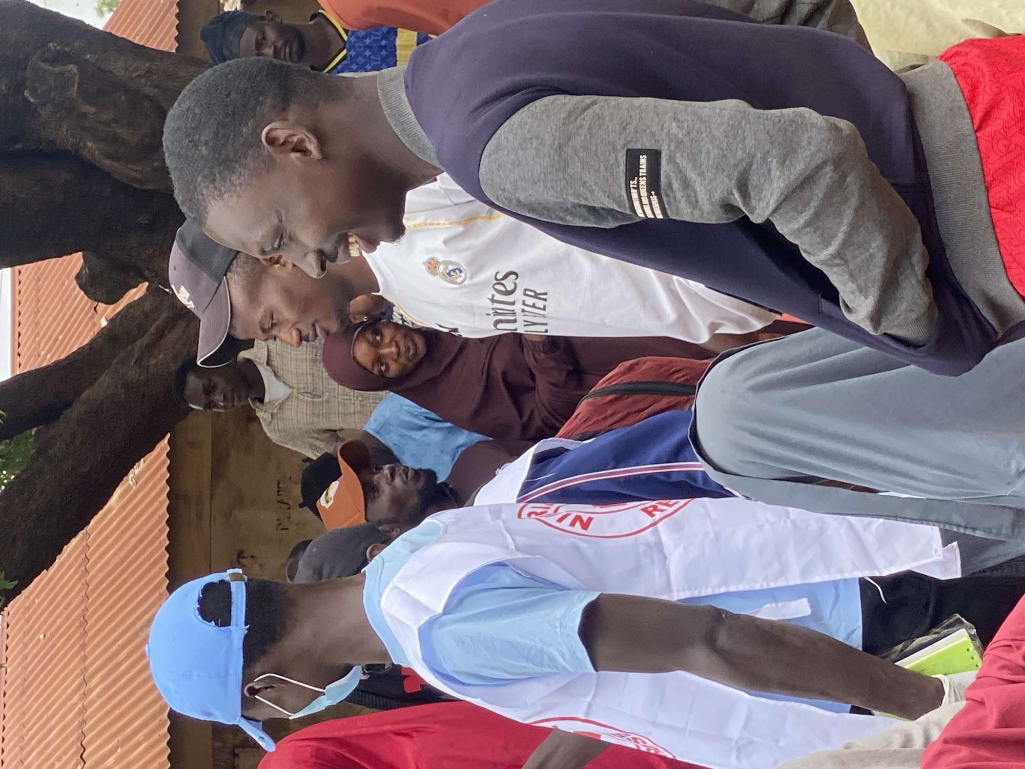 This media file shows a group of people, predominantly young men, gathered outdoors. The scene appears to be in a rural or semi-urban setting, possibly in Rigachikun, Nigeria, given the context. The background features a corrugated metal roof, suggesting simple structures, and a large tree trunk providing shade. The lighting suggests it is daytime, likely overcast or late afternoon, as there are no harsh shadows.
The individuals are dressed in a variety of casual clothing. One person in the foreground wears a blue baseball cap and a light blue t-shirt, with a white and red vest over it. This vest has visible text, appearing to be "RE" and possibly other letters in red on a white background, perhaps part of a logo or slogan. Another individual is wearing a white jersey with "R. Madrid" and a crest visible, resembling a football club's kit. Further back, other people are seen wearing t-shirts, caps, and some form of head covering.
The people seem to be interacting or observing something, with many looking in the same direction, though their expressions are not entirely clear due to the angle and focus. The overall atmosphere seems informal, perhaps a community gathering, a sporting event, or a casual meet-up. One person in the center is captured mid-speech or laughter, showing teeth and a lively expression. The presence of a face mask on one individual suggests ongoing health precautions. The ground is not clearly visible, but the overall impression is of an outdoor, community-oriented gathering.