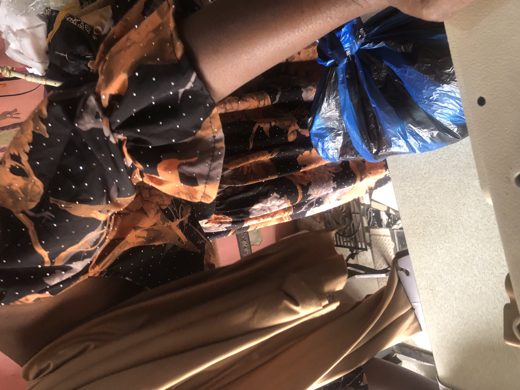 The image depicts a close-up view of fabric and a person's arm, suggesting a setting possibly related to tailoring or clothing production in Ado, Nigeria. A tan-colored fabric hangs prominently on the left, showing folds and a smooth texture. To the right, a black fabric with an orange, possibly floral or abstract pattern, and scattered white dots is visible. The sleeve of a garment made from this patterned fabric is also shown, revealing a person's arm with visible pores. In the foreground, a crumpled blue plastic bag, partially tied, rests on a light-colored surface. The background is out of focus, but hints of a colorful wall and what appear to be garments or textiles on hangers can be discerned. The lighting suggests an indoor environment.