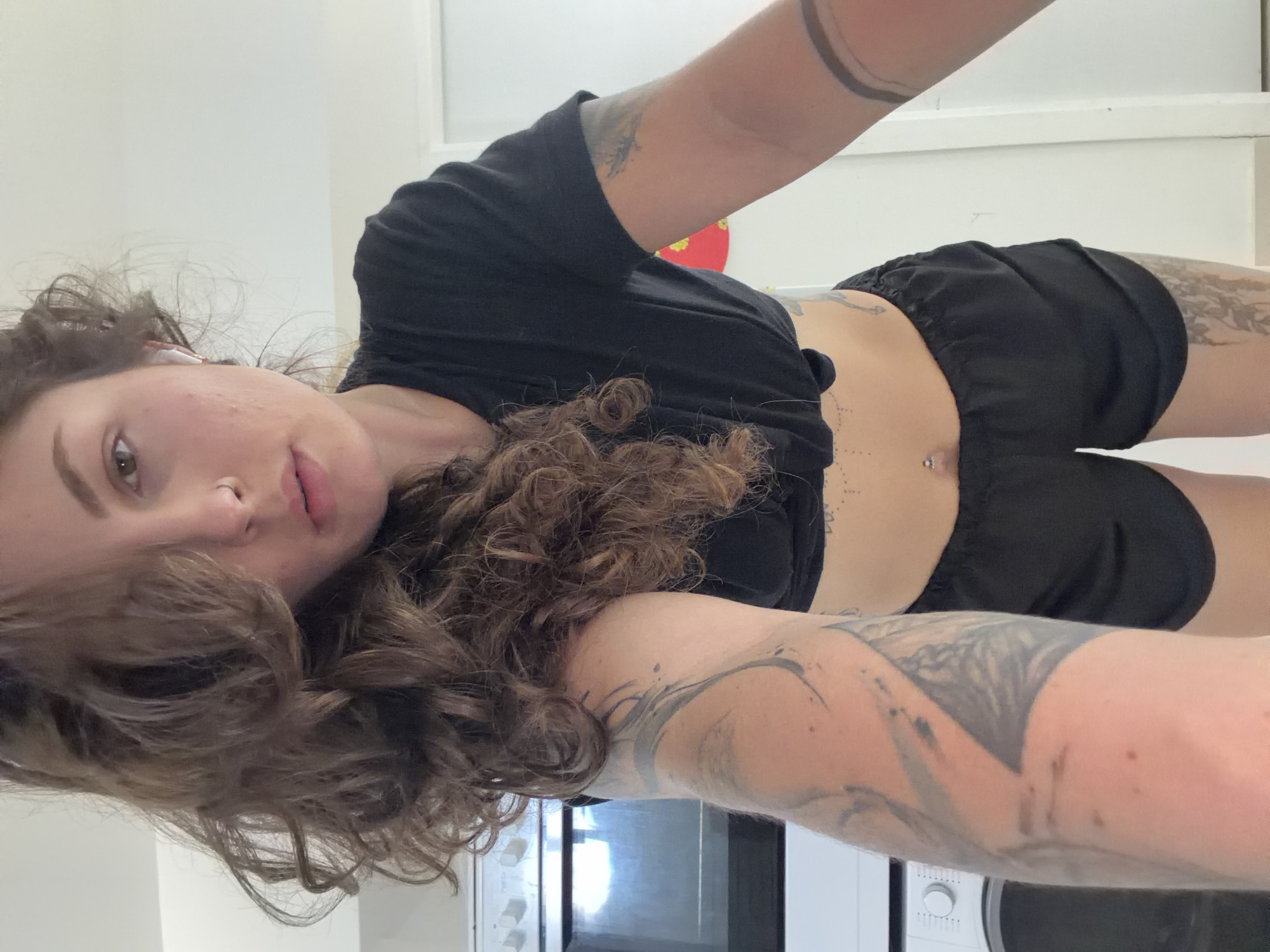 A person with medium-brown, curly hair is featured in a close-up, rotated selfie, appearing to be lying down or leaning back indoors. The individual, likely female, is looking directly at the camera with a neutral expression. An earbud is visible in her right ear.
She is wearing a black crop top that exposes her midriff and black shorts. Distinctive body modifications include a nose piercing (ring) and a navel piercing. Extensive tattoos are visible on both arms, the side of her torso, and a dotted pattern around her navel. Floral or nature-inspired tattoos are also visible on her legs/thighs.
The background consists of plain white walls, suggesting a minimalist indoor setting. Elements of a kitchen, possibly an oven or stovetop, are faintly visible in the lower portion of the image. A round, red object with yellow details, possibly a decorative item, is partially visible in the upper background. The lighting is even, typical of indoor conditions. No visible text is present.
The scene is set in Kiryat Bialik, Israel.