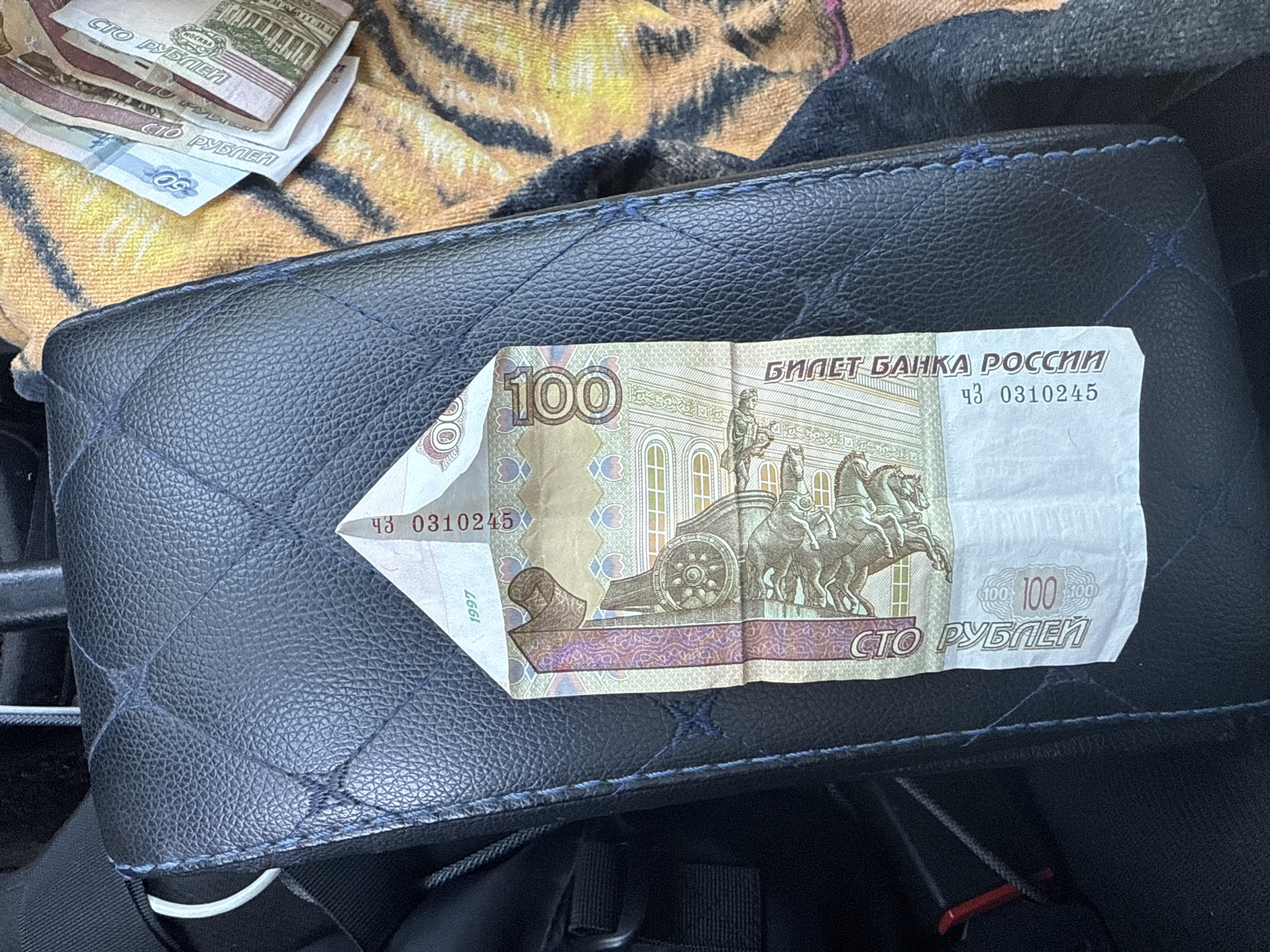 A folded 100-ruble banknote from Russia is placed on a black quilted surface, possibly an armrest or car seat. The banknote is creased in the center, forming a triangular shape at the top. The denomination "100" is clearly visible on the banknote, along with the text "СТО РУБЛЕЙ" (One Hundred Rubles) and "БИЛЕТ БАНКА РОССИИ" (Banknote of the Bank of Russia). A serial number "43 0310245" and the year "1997" are also visible. To the right of the black surface, a fabric with a tiger-striped pattern is partially visible. The image is taken from a slightly elevated angle. There is no information available to determine the specific location in the city of Samara, Russia.