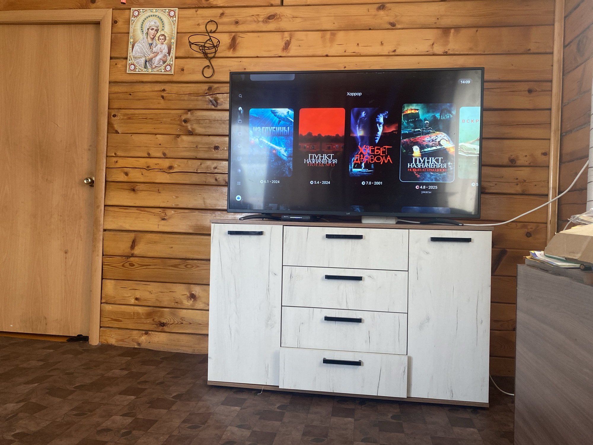 The image shows a television screen displaying movie posters in a room with wooden paneled walls. A door is visible on the left side of the image, and a white dresser with drawers is positioned beneath the television. Above the television, an icon of the Virgin Mary and child is mounted on the wall. The scene is indoors, and the overall aesthetic suggests a rustic or cabin-like interior, which aligns with the provided location information of the village of Kulchum, Russia.