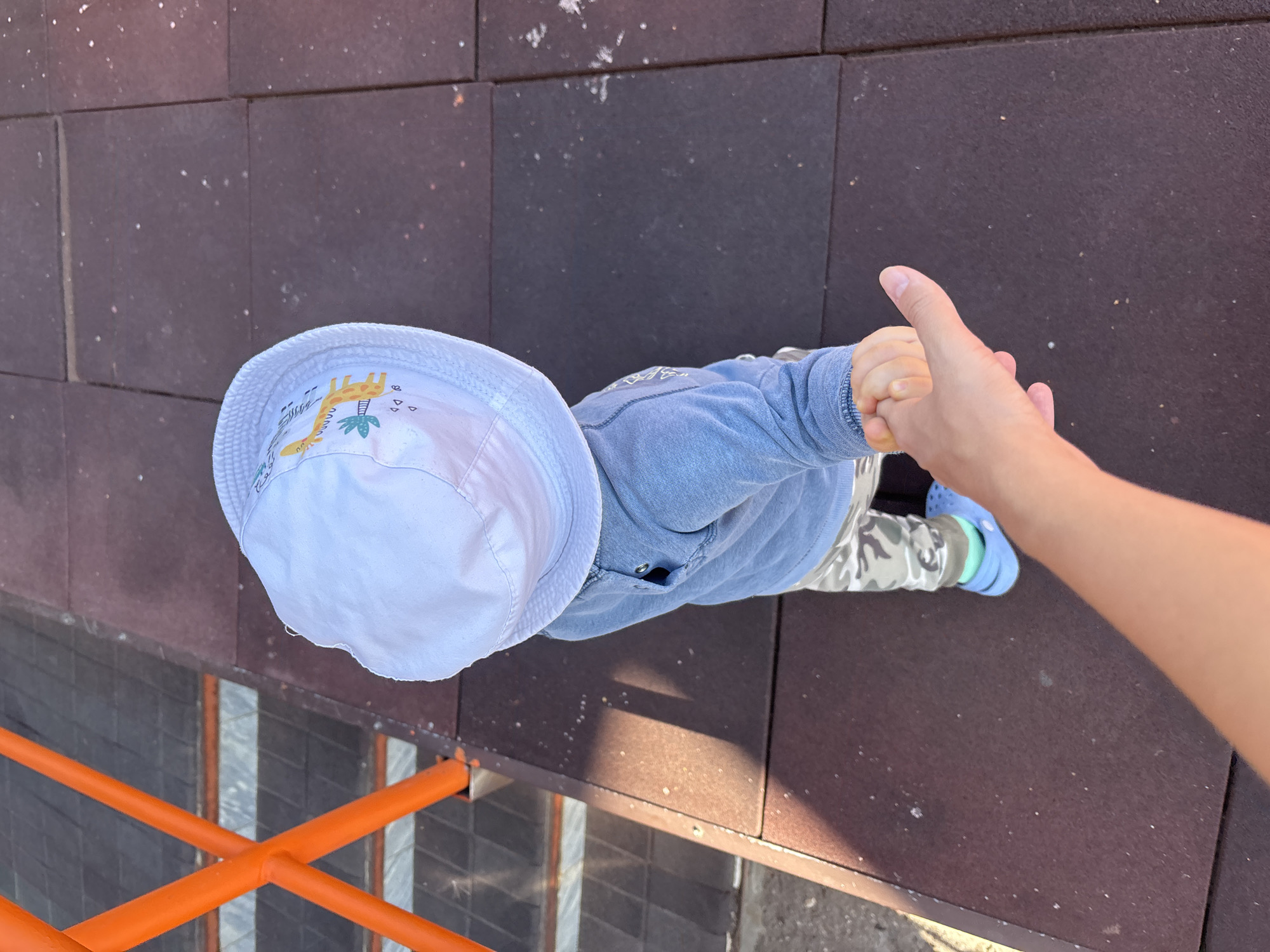 An overhead perspective captures a toddler wearing a light blue long-sleeved shirt, camouflage pants, a white sun hat with a giraffe print, and blue Crocs, holding an adult's hand. They are standing on a dark brown rubberized playground surface, with orange metal railings and grey steps of a playground structure visible to the left. The scene appears to be a section of a playground in Naberezhnye Chelny, Russia, likely during daylight hours given the shadows.
