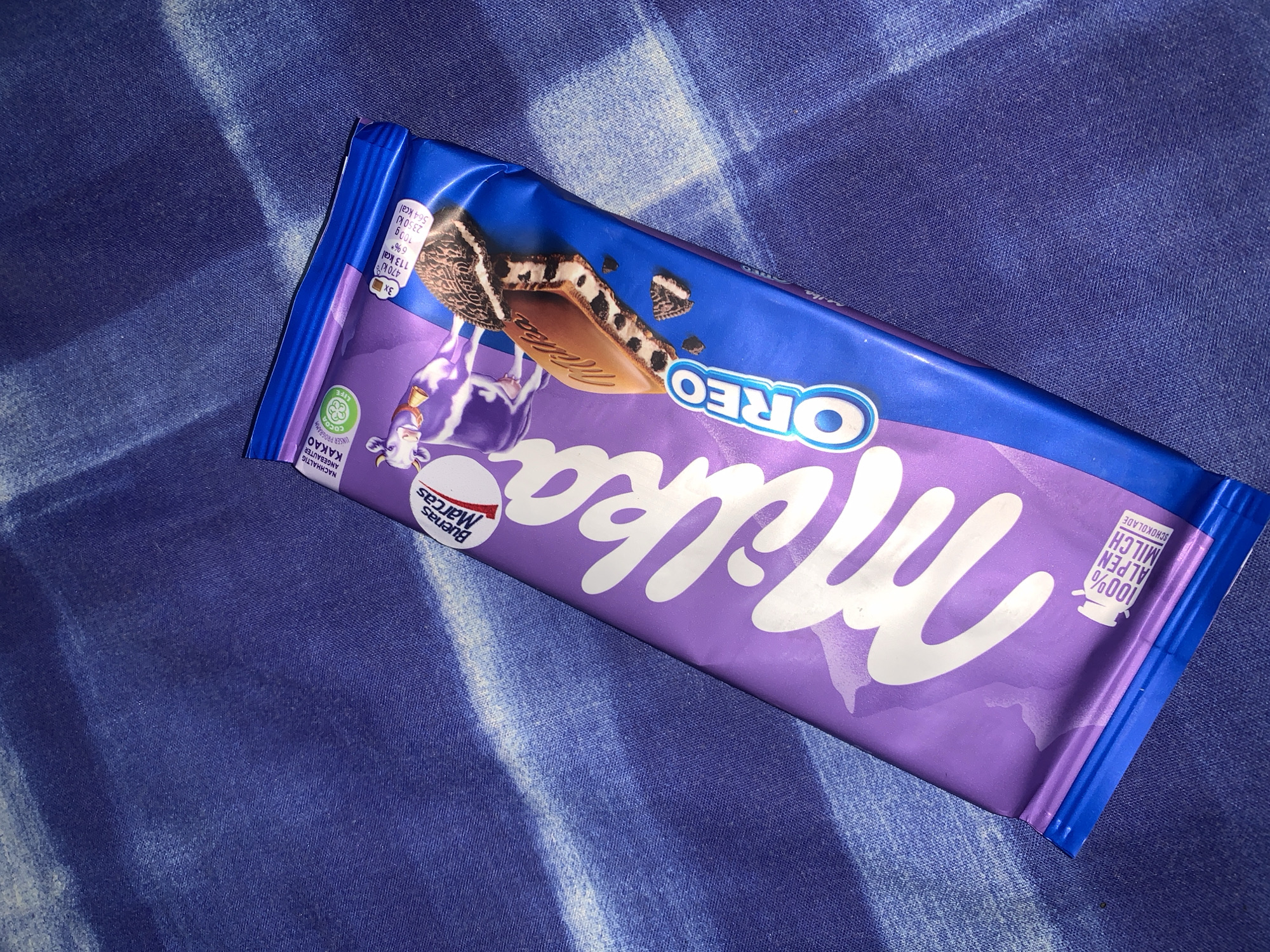 A Milka Oreo chocolate bar wrapper lies diagonally on a blue striped fabric surface. The wrapper is predominantly purple and blue with white lettering and images. The Milka logo is prominently displayed in white. The Oreo logo is also visible in white. The wrapper also includes nutrition information and branding related to sustainable cocoa production. The fabric beneath the wrapper has a distinct blue and white striped pattern. There is no indication of the city of Santa Ana, Venezuela or any people in the image.
