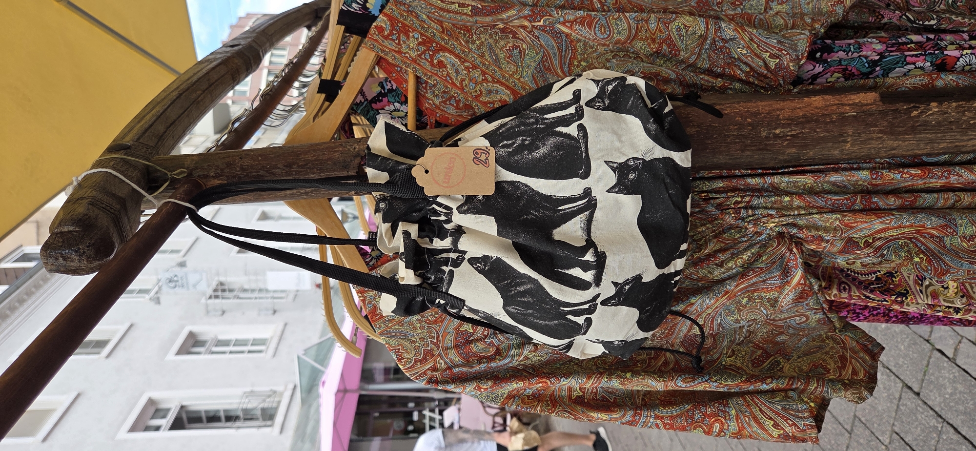 A drawstring bag with a black and white cat print is displayed on a wooden stand, amidst clothing items hung on hangers. The bag has a small brown tag attached, showing the word "tumico" in pink lettering and the number "29.". The background features a street scene with buildings, a yellow umbrella, and a pink awning. The scene appears to be an outdoor market stall. The location is identified as Karlsruhe, Germany, likely at a street market.
