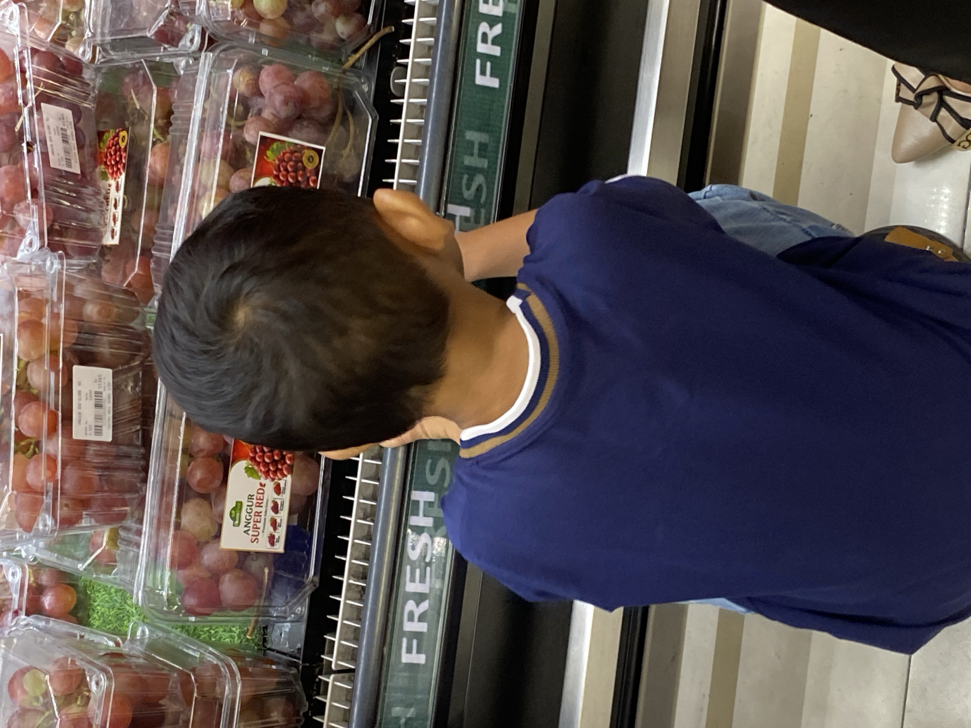 The image shows the back of a young boy in a dark blue shirt with striped trim, looking at a display of red grapes in plastic containers at a supermarket. The grapes are stacked in clear plastic packaging, with labels indicating they are "ANGGUR SUPER RED." In the background, a green sign with the word "FRESH" is visible. The setting appears to be a grocery store aisle, likely in Banda Aceh, Indonesia, given the label text.