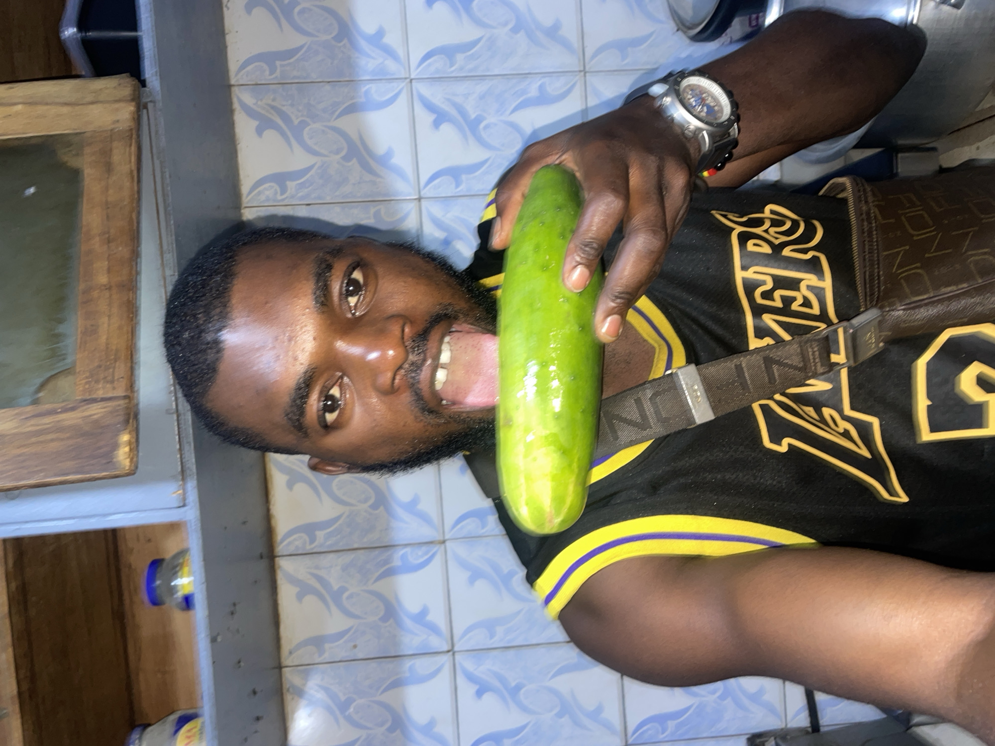 A man stands in a room with blue and white patterned tile walls. He is holding a cucumber in front of his mouth. He is wearing a black Lakers jersey with yellow trim, a brown patterned satchel, and a wristwatch. A wooden cabinet is visible in the background, with water bottles inside. The specific location within Jos, Nigeria is indiscernible from the image.
