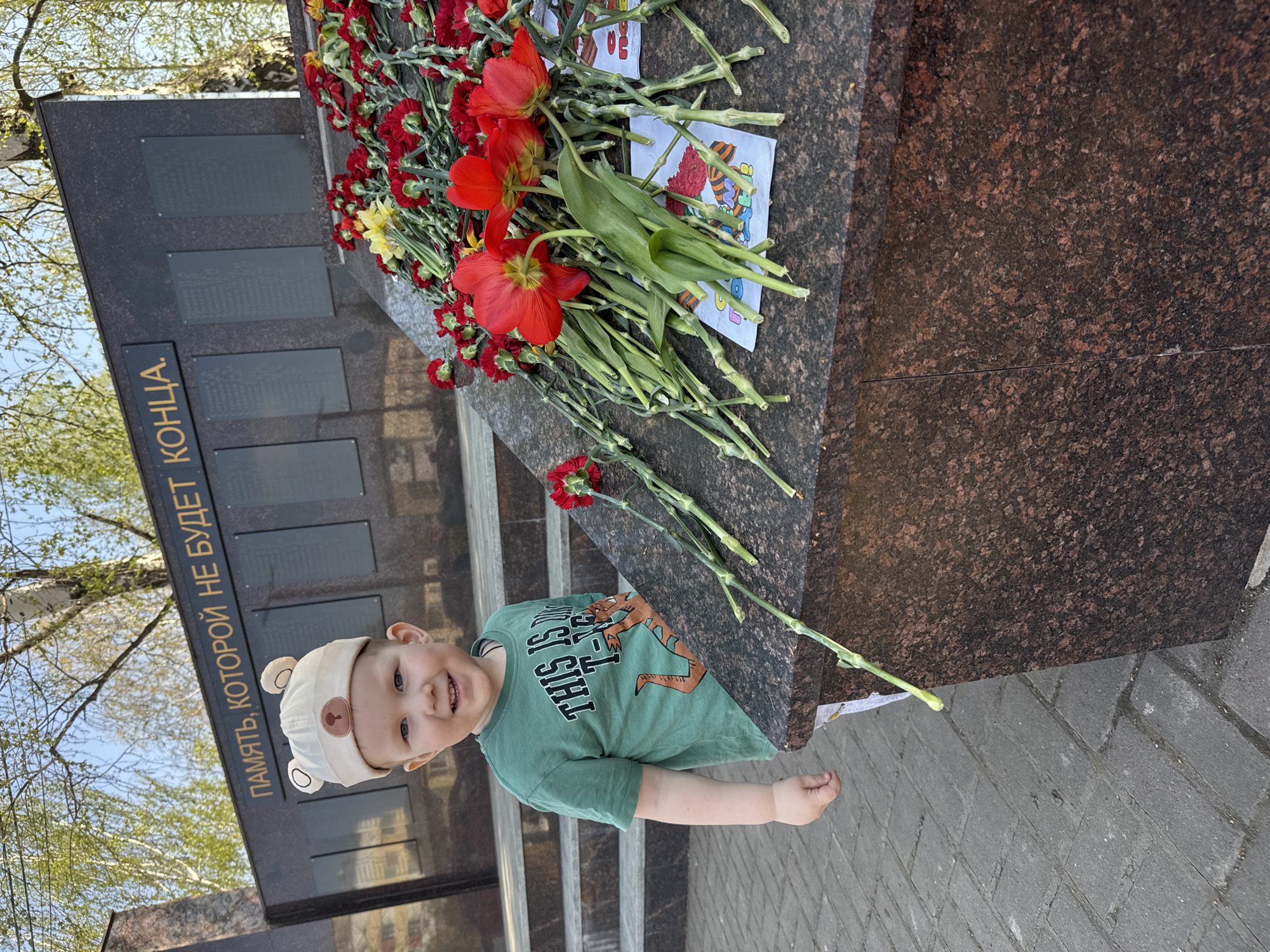 A young child wearing a bear-themed hat and a green t-shirt stands in front of a granite monument adorned with red and yellow flowers. The monument has multiple plaques with Cyrillic writing. The inscription on the upper plaque reads "ПАМЯТЬ, КОТОРОЙ НЕ БУДЕТ КОНЦА". The ground is paved with gray bricks. The location is identified as Zav'yalovo, Russia.