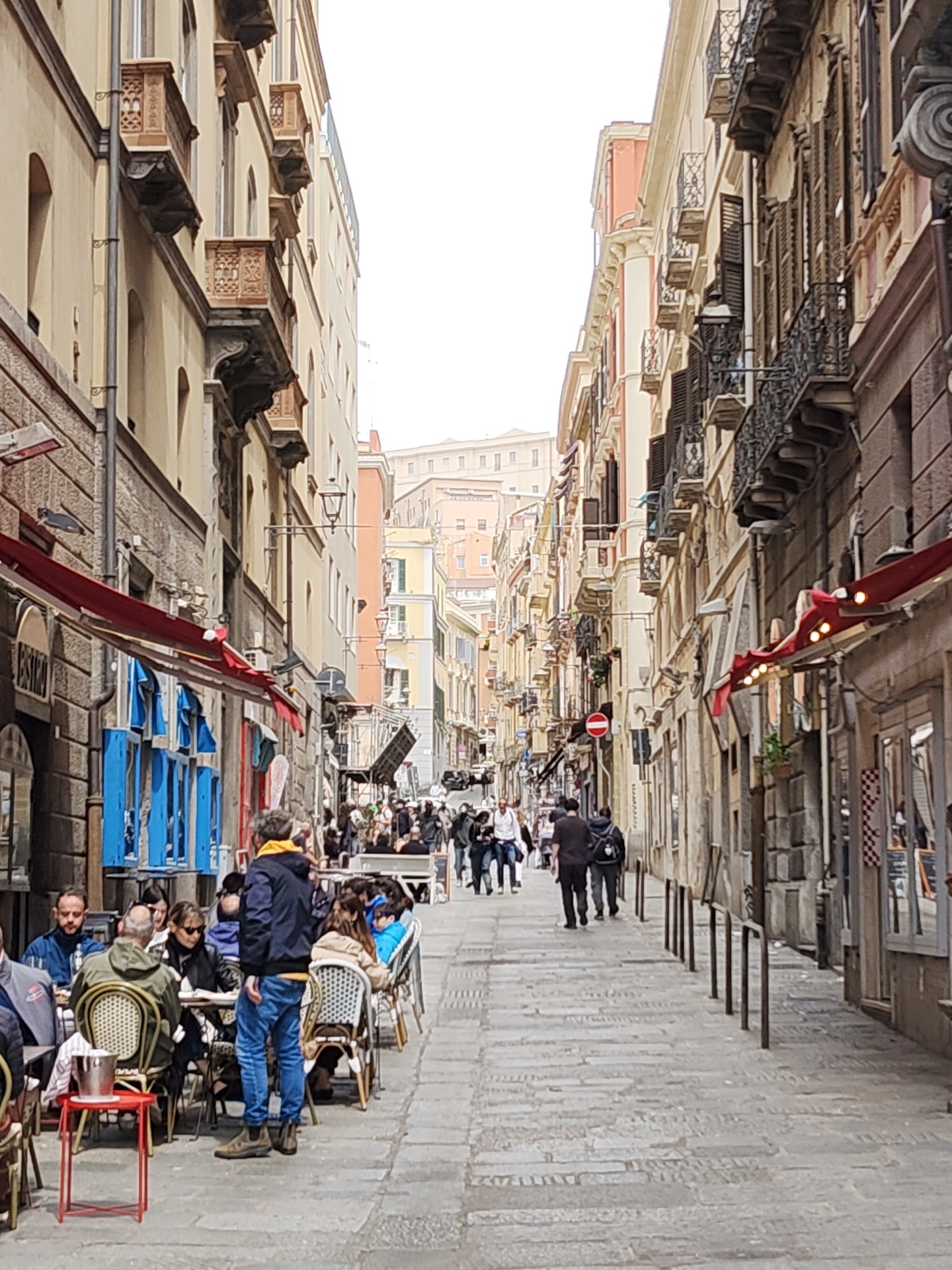 The image depicts a narrow street in Cagliari, Italy, populated with people and lined with buildings. The street is paved with stone blocks. On the left side, there is a restaurant with outdoor seating where people are seated at tables, eating and talking. The buildings on both sides of the street are multi-storied with balconies and various architectural details, including shutters and awnings. A group of people is walking further down the street, which narrows into the distance. A red "no entry" sign is visible on the right side of the street. The sky is overcast, casting a soft light on the scene.