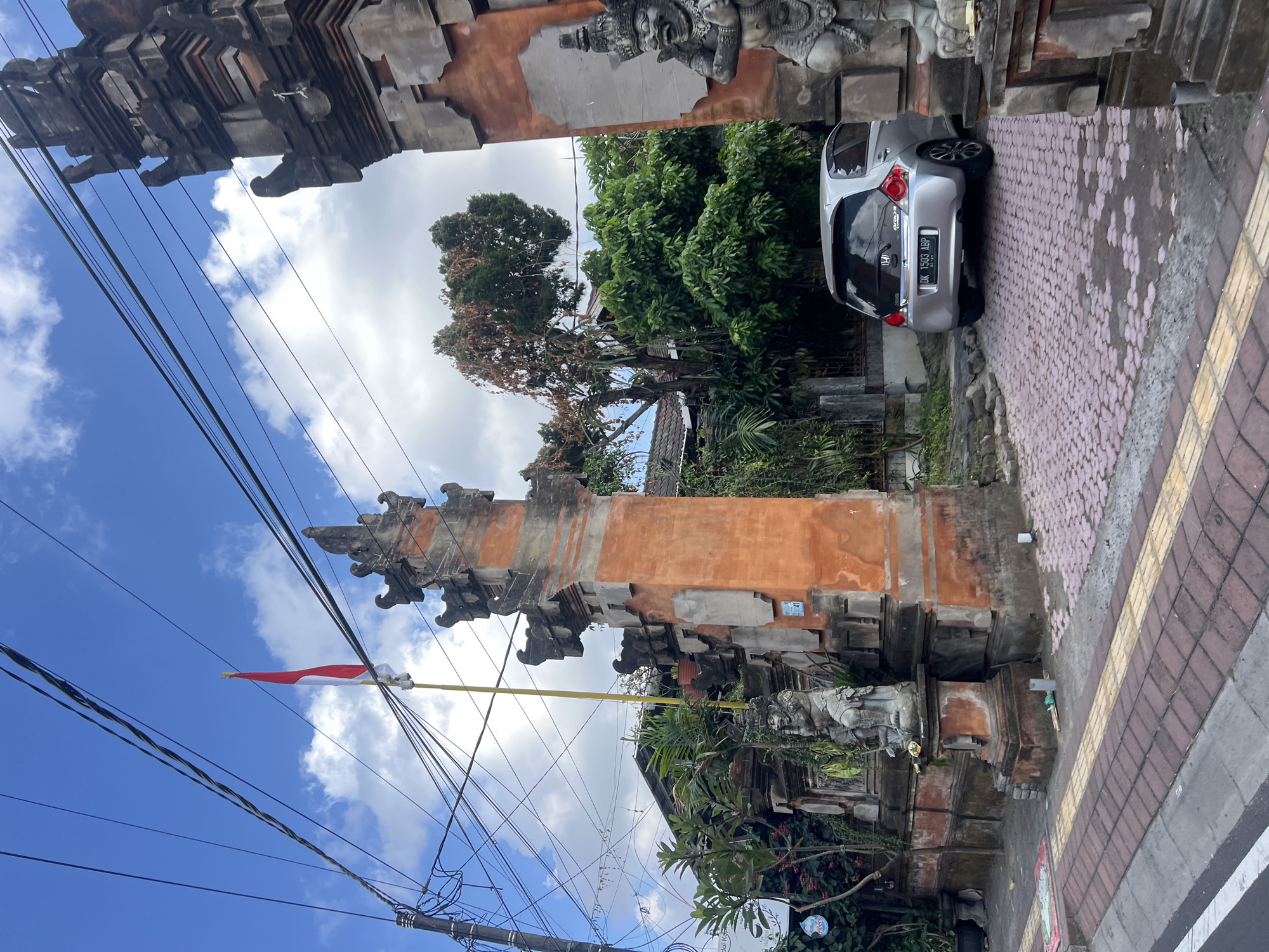 An outdoor, eye-level, vertical shot captures a scene in Denpasar, Indonesia, on a partly cloudy day. The foreground shows a sidewalk with red and gray paving stones, leading to a street. A traditional Balinese stone gate, adorned with ornate carvings and a red and orange-hued plaster, stands prominently in the center-right of the frame. The gate is flanked by two imposing stone guardian statues.
Behind the gate, a silver Honda Brio car is parked on a driveway paved with pink hexagonal stones. The car's license plate reads "DK 1503 ABP". Lush green vegetation and trees surround the gate and extend into the background, where a glimpse of a tiled roof residential building is visible. A tall flagpole with the Indonesian flag, red over white, stands to the left of the main gate structure, silhouetted against the bright blue sky dotted with white clouds. Numerous electrical wires crisscross the sky above the scene, typical of urban environments. The overall impression is one of cultural heritage meeting modern daily life in a tropical setting.